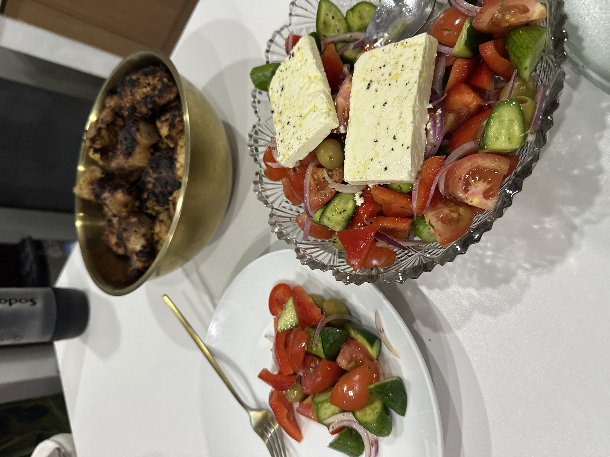 This image depicts a meal, possibly in North Kuta, Indonesia, consisting of a Greek salad and grilled meat. The Greek salad, featuring cucumbers, tomatoes, red onions, green olives, and feta cheese, is presented both in a decorative glass bowl and on a white plate, accompanied by a gold fork. A gold bowl containing grilled meat is also visible. A soda siphon is present in the scene.