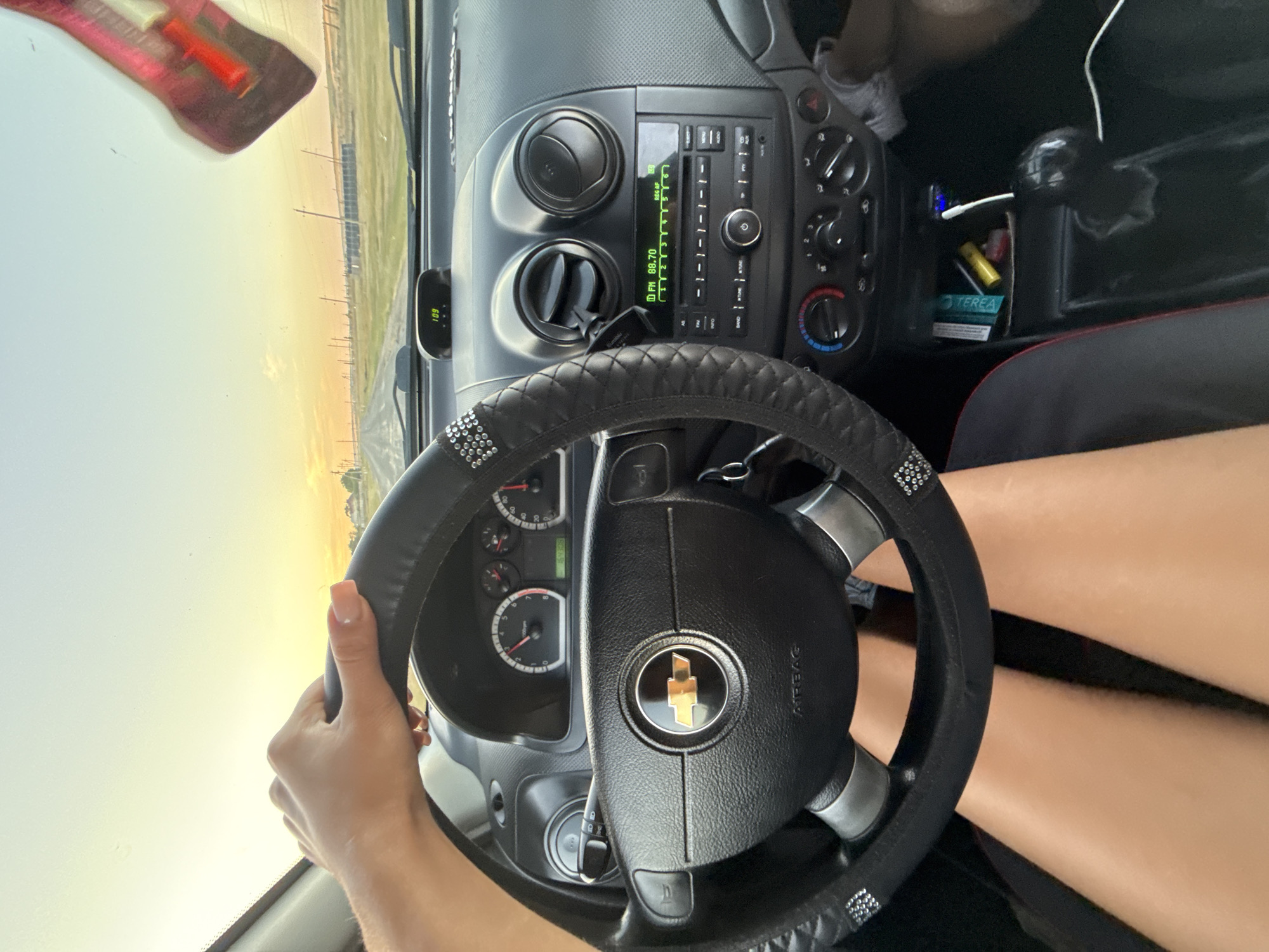 The image depicts the interior of a car from the driver's perspective, with the sunset visible through the windshield. A hand, presumably the driver's, rests on the steering wheel. The dashboard features a radio, air vents, and various controls. A digital clock on the dashboard displays "109", and the radio is tuned to "FM 88.70". Various items, including what appears to be a pack of cigarettes and a yellow lighter, are visible in a compartment below the radio. A pink, guitar-shaped air freshener hangs from the rearview mirror. The scene is set on a road in Cioc-Maidan, Moldova, with utility poles and fields visible in the distance. The image captures a moment of travel during a sunset.