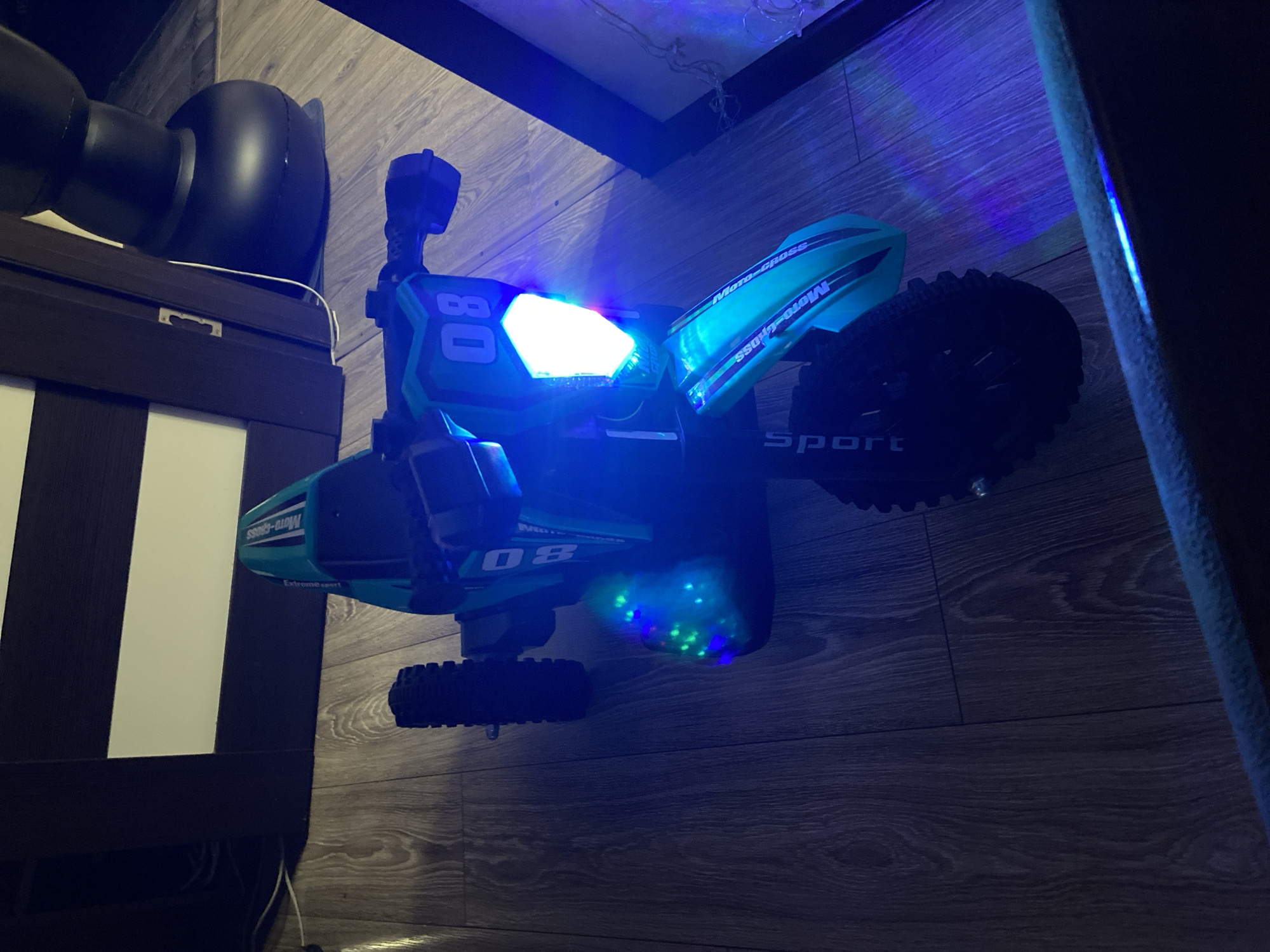 A toy motocross bike with blue and green lights is displayed on a wooden floor. The bike is predominantly teal and black with "Moto-Cross" written on the fenders and "Sport" on the rear wheel guard. The number "08" is visible on the front of the bike, next to a bright white headlight. Several green and blue LED lights are scattered across the underside of the bike, illuminating the floor beneath it. The scene is dimly lit, with the bike's lights being the primary light source, casting blue light onto the surrounding wooden floor and a wall. The background includes a dark brown furniture piece with vertical white stripes and part of a large black object, possibly a speaker or furniture, in the upper left corner. The overall impression is that of a staged display or a close-up product shot in a domestic setting in Beloretsk, Russia. There is no indication of time of day or weather.