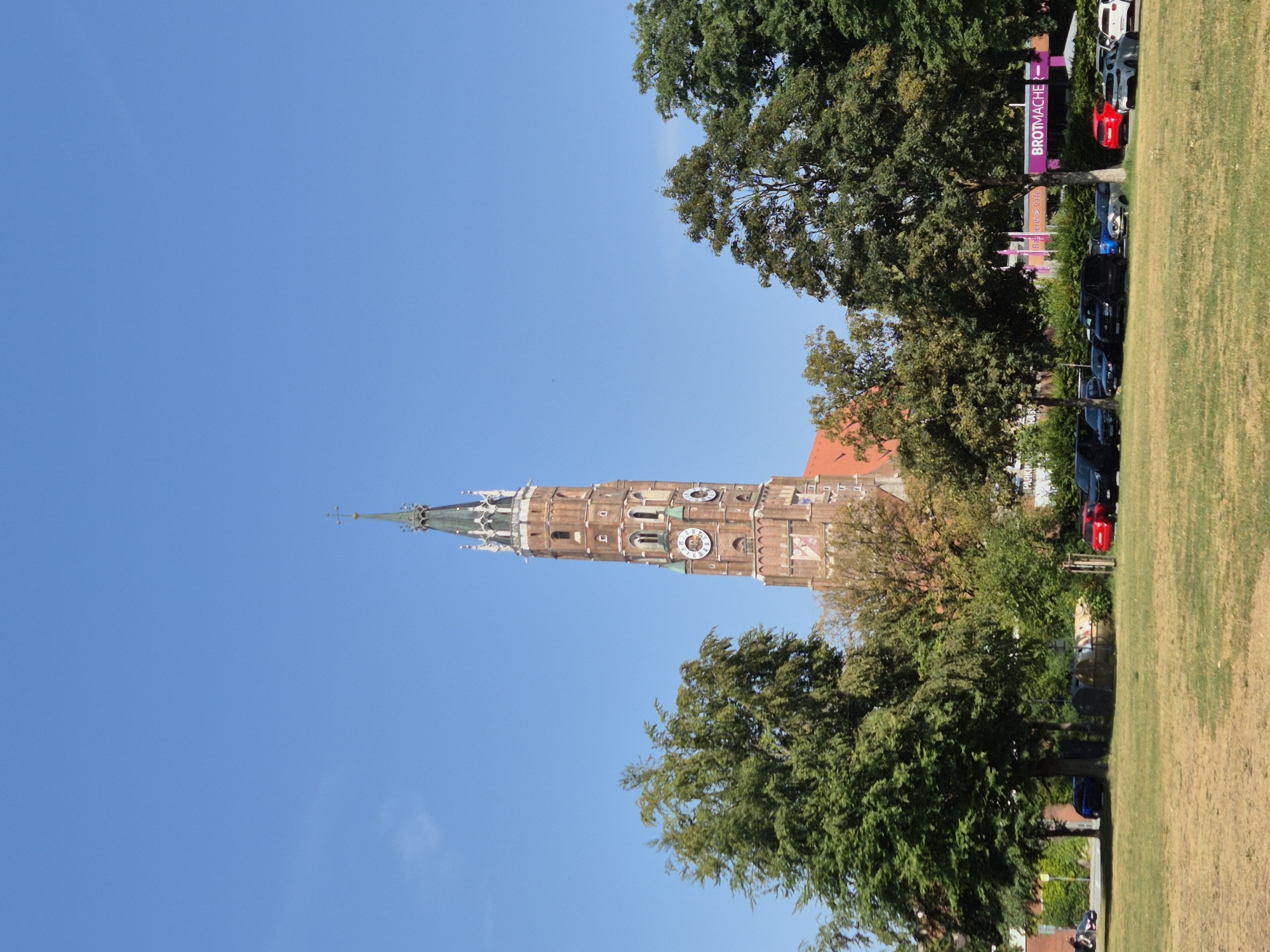 The image shows the St. Martin's Church tower in Landshut, Germany, dominating the skyline under a clear blue sky. The Gothic brick tower, a prominent landmark of the city, features clocks and intricate architectural details. In the foreground, a grassy field transitions to a line of trees and a parking lot filled with various cars, including red, black, and silver vehicles. A sign with the text "BROTMACHER" and "Bäckerei > Cafe" is visible, indicating a bakery and cafe. A person on a scooter is seen on the left side of the image, and several trash bins are located near the trees. The scene depicts a sunny day in an urban environment, with the church as the central architectural element.