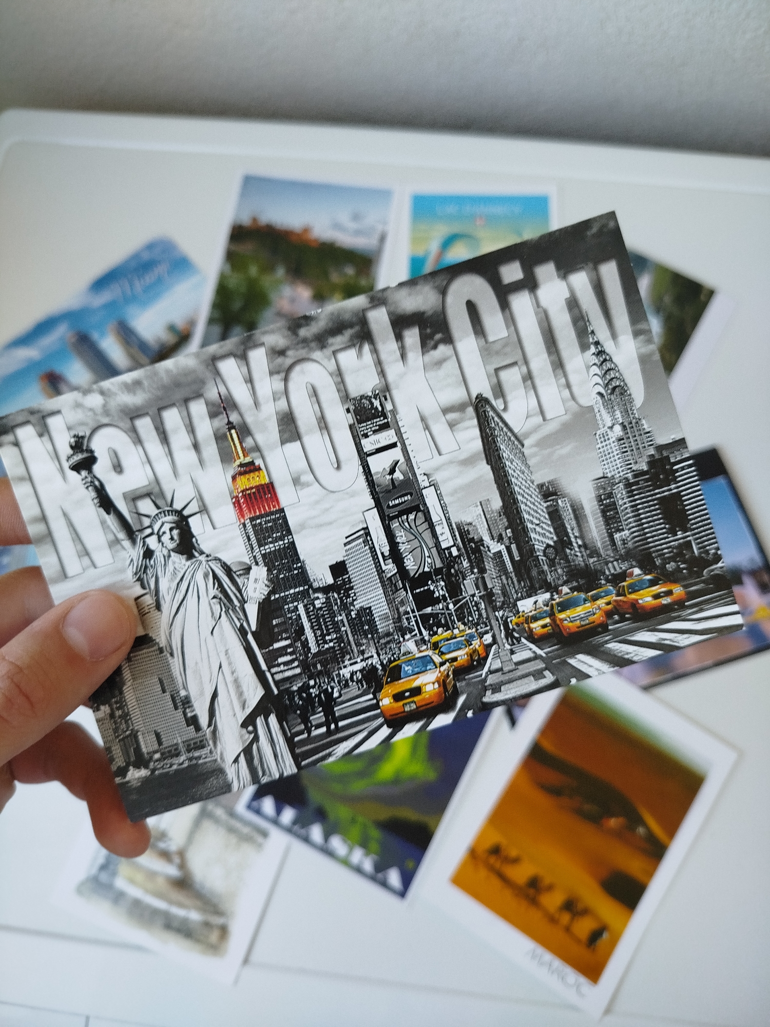 A hand holds a postcard depicting a black and white photograph of New York City, with the Statue of Liberty, the Empire State Building (partially colored red and gold), Times Square, the Flatiron Building, and the Chrysler Building. Yellow taxis are seen on the street. Surrounding the NYC postcard are other postcards, including one that reads "ALASKA" and another "MAROC" (Morocco). The scene is set on a white surface. There is no indication of the city of Varna, Bulgaria in the image.