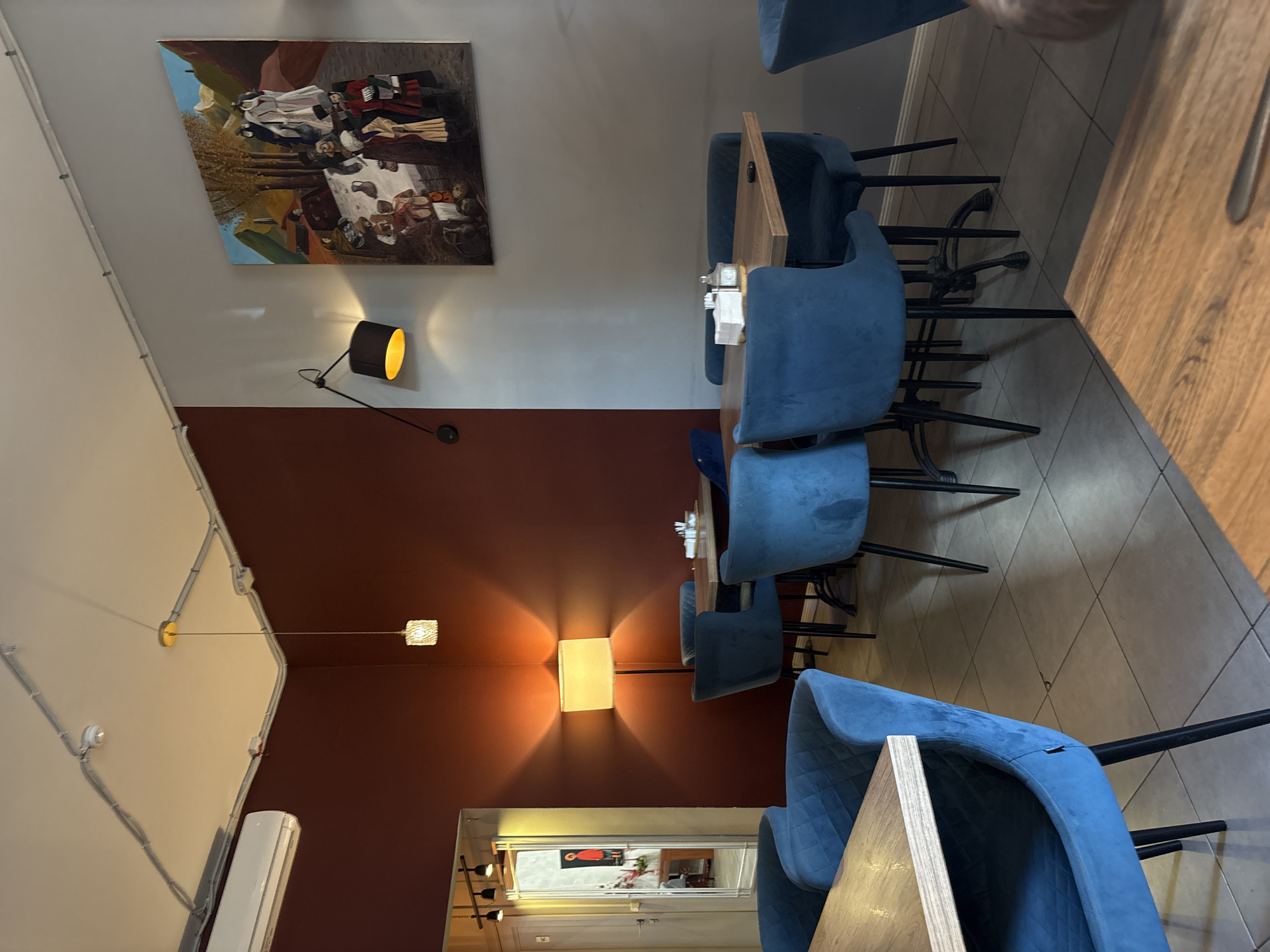 The image shows a dimly lit interior of a restaurant or cafe. The scene is dominated by blue upholstered chairs and wooden tables. In the background, a painting hangs on a light-colored wall, depicting figures in what appears to be a historical or allegorical scene. Several lamps are visible, including a black articulated lamp with a bright orange light source, and a rectangular lampshade casting a warm glow on a reddish-brown wall. Another smaller lamp with a textured shade hangs from the ceiling. The flooring is tiled in a diamond pattern. The overall impression is one of a cozy and somewhat intimate setting, perhaps in the evening or with subdued lighting. There are no people visible in the immediate scene, suggesting it might be closed or empty at the time the photo was taken. The lighting creates a somewhat moody atmosphere.