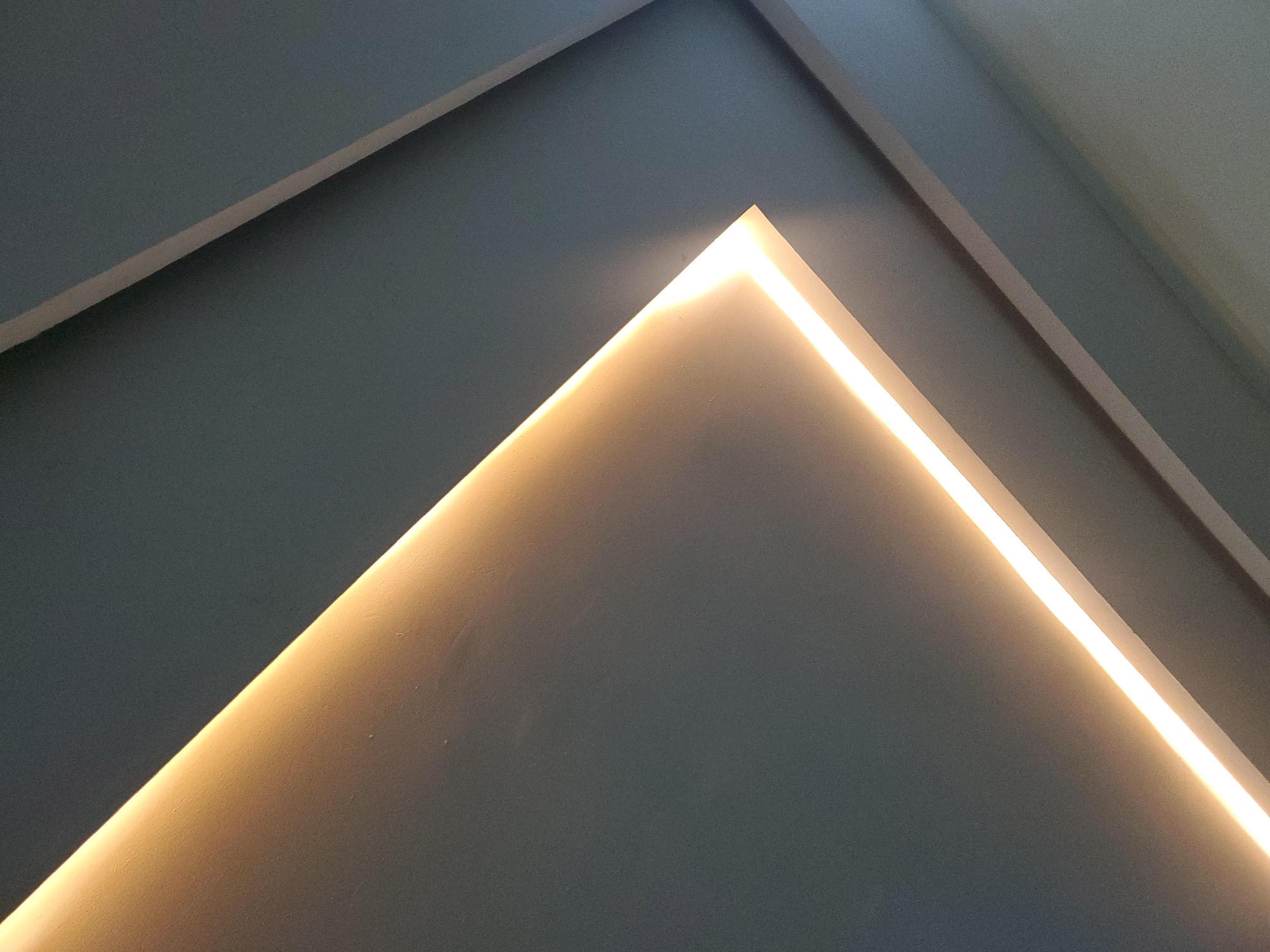 This is a close-up, abstract photograph of architectural lighting. The primary subject is a recessed light fixture emitting a warm, golden glow. The light creates a sharp, triangular shape against a dark grey, textured surface, likely a wall or ceiling. The light itself is a bright, continuous line, creating dramatic shadows and highlights on the surrounding architecture.
The scene is geometric and minimalist, with clean lines and subtle gradients of light and shadow. The texture of the surface is visible, suggesting a painted or plastered finish. The overall impression is one of modern interior design, possibly in a contemporary building.
There is no discernible activity or event taking place, as the image focuses solely on the static architectural elements. There are no people, animals, or objects other than the lighting and the building materials.
Notable details include the stark contrast between the bright light and the dark surfaces, and the way the light emphasizes the angles and planes of the architecture. The time of day and weather are not discernible, and there are no specific location cues beyond the general architectural style. The mood is one of calm and sophistication, with a focus on light and form. No text is visible in the image. The context provided, Jos, Nigeria, suggests this is likely an indoor setting within that location.