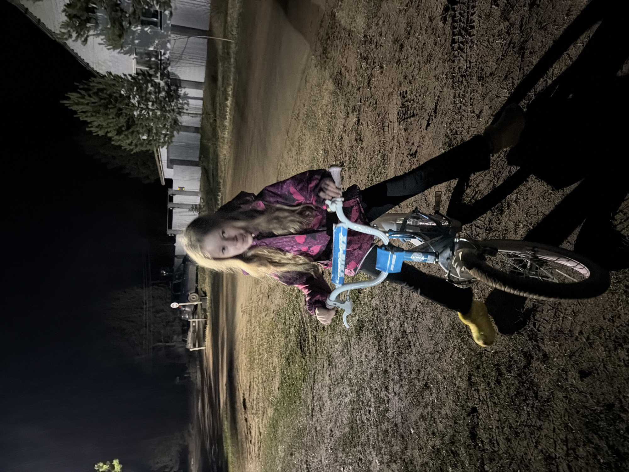 The photograph shows a young person on a bicycle at night. The individual is wearing a dark-colored hooded jacket with a pink and magenta pattern, dark leggings, and light yellow shoes. They are holding the handlebars of the bicycle, which has a blue frame. The setting appears to be a dirt or gravel area, possibly a roadside. In the background, there are buildings or structures, and trees are visible on the left side of the frame. The left of the frame is mostly dark, contrasting with the relatively brighter area where the individual and bicycle are located. The image is slightly rotated clockwise. The location in the city of Markovo, Russia is not discernible from the content of the provided image.
