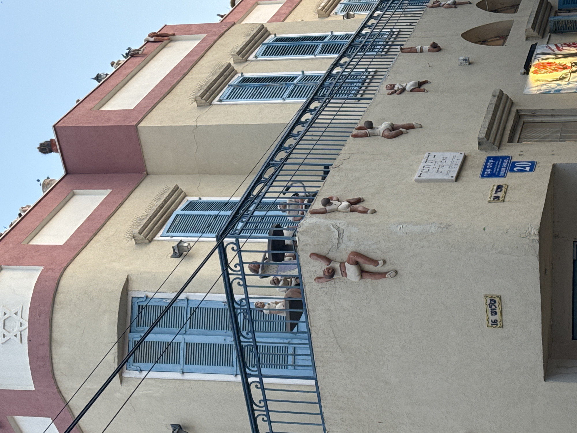 The image captures a multi-story building facade in Tel Aviv, Israel, under a clear, bright daytime sky. The building is primarily light beige with reddish-brown and white trim, featuring several windows with distinct blue louvered shutters.
A prominent and unique artistic installation adorns the building's exterior: numerous small, stylized human figures, resembling children, are attached to the walls in various climbing, crawling, and hanging poses, creating a whimsical effect. A dark blue metal railing, possibly part of a fire escape or decorative element, runs diagonally across the facade.
Along the roofline and ledges, several pigeons are perched. On the left side of the building, a carved Star of David emblem is visible. Black electrical wires traverse the scene.
Visible text elements include a blue street sign displaying "שלום שבזי" (Shalom Shabazi), a street name in Hebrew, and the number "20." Below it, a smaller yellow and blue ceramic plaque also shows Hebrew text and the number "16." A white rectangular plaque with additional Hebrew script is mounted on the wall. A colorful artwork or mural is partially visible near an upper window.