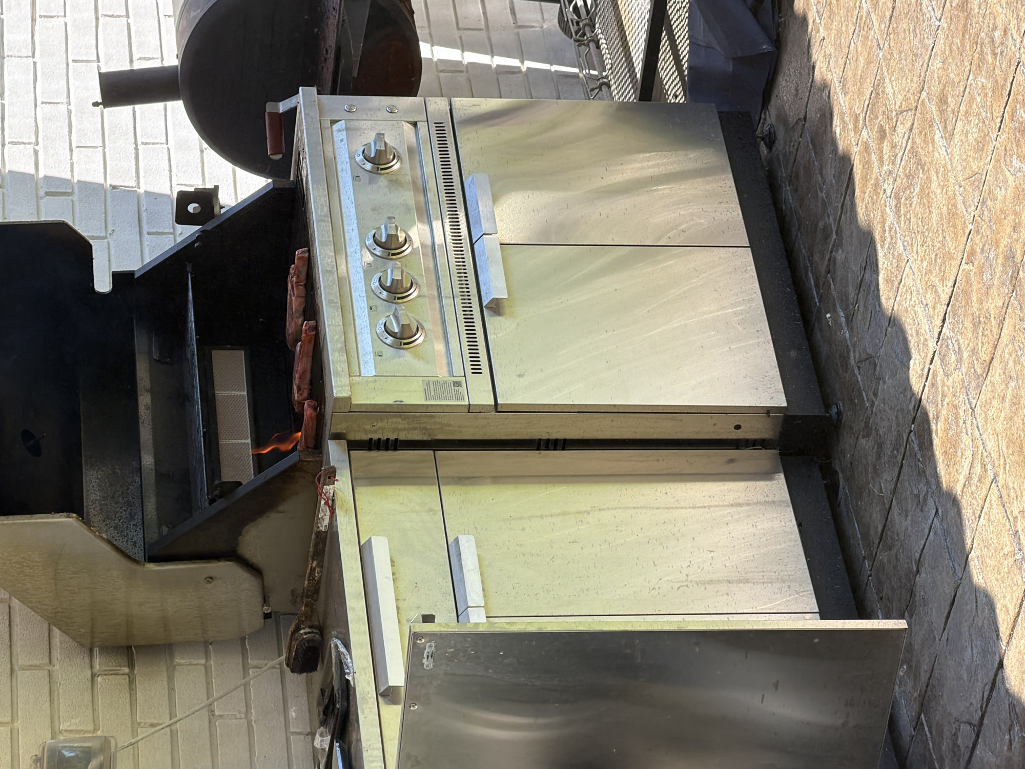 An outdoor grilling area is depicted in Pepper Pike, United States of America. The central feature is a stainless steel outdoor kitchen unit incorporating a gas grill with an open lid. Several cuts of meat, including steaks and hot dogs, are visible cooking on the grill grates, with active flames emanating from below. The grill's front panel contains four control knobs and a horizontal ventilation grate. Below the grill, two stainless steel cabinet doors with horizontal bar handles are present. To the immediate left, an additional stainless steel cabinet unit with a corresponding handle extends the setup. A long-handled grill brush and other small items rest on the countertop adjacent to the grill. The background features a white-painted brick wall. Partially visible to the right against the wall is a large, dark, barrel-style smoker or grill equipped with a side handle and a vertical exhaust stack. The ground is paved with brown and tan stamped concrete, exhibiting strong, elongated shadows indicative of direct sunlight. No individuals are present within the scene.