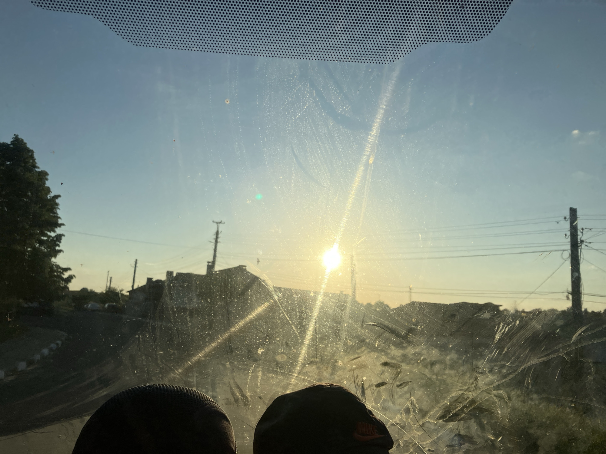 The image shows a view through a dirty vehicle windshield. The location appears to be Komuniga, Bulgaria, judging by the architecture and utilities present. In the distance, there is a residential area with houses and utility poles. The sun is shining brightly, causing glare and obscuring details. The foreground shows two dark, possibly baseball, caps resting on the vehicle's dashboard.