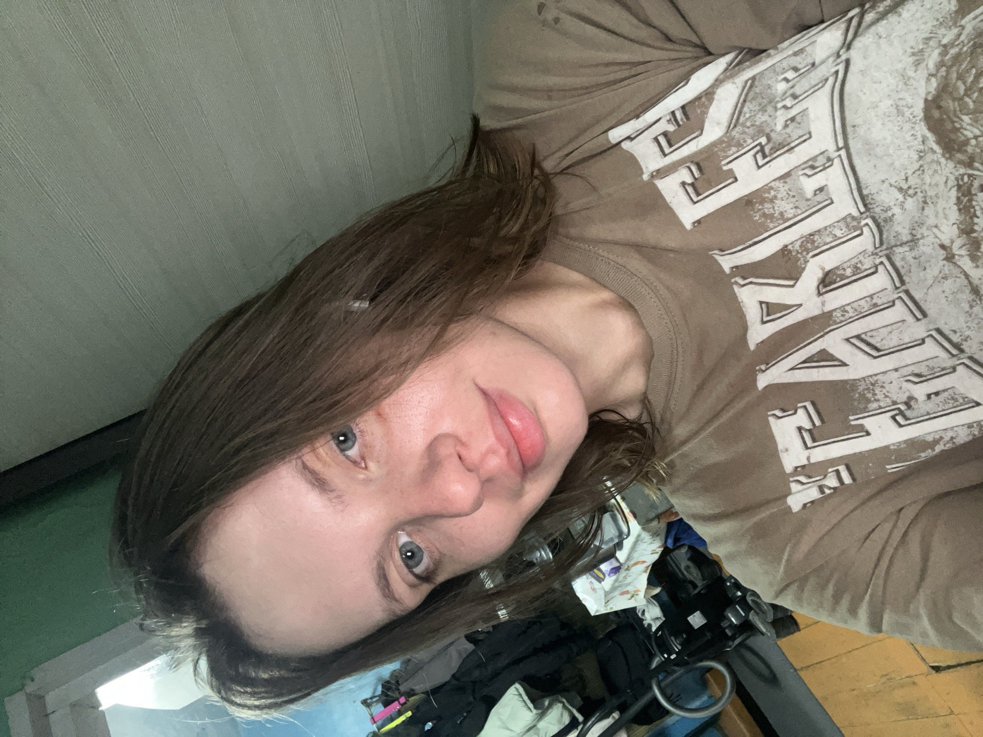 A young Caucasian woman with medium brown hair and blue eyes is taking a selfie in an indoor environment. She is wearing a light brown t-shirt with distressed white lettering that appears to say "FEARLESS". Her skin has a light dusting of freckles.
The background shows a room with green painted walls and a wooden floor. To the left, there is a rack of clothing, including a white puffy jacket and dark garments. A table covered with a patterned cloth is visible behind her. The overall impression is a casual, personal space. There is no indication of the specific location within the city of Nizhny Tagil, Russia.