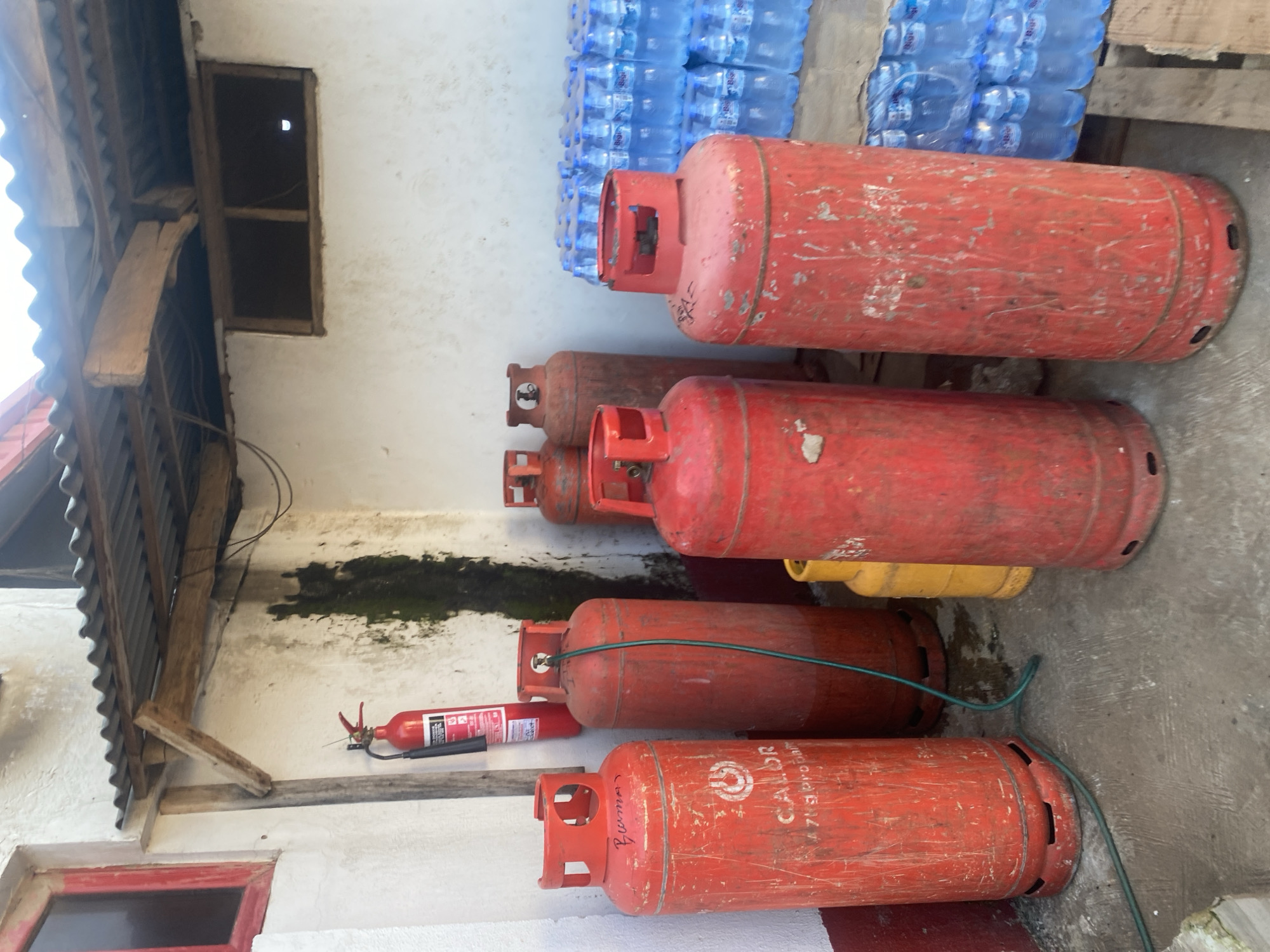 In a cluttered outdoor storage area, likely behind a building in Ikorodu, Nigeria, several red propane gas cylinders are arranged. One cylinder, on the far left, has "CALOR" written on it. A green hose connects a smaller cylinder to a larger one. To the right of the cylinders, stacks of bottled water are visible. A red fire extinguisher hangs on a wall behind the cylinders. The ground is concrete, and a corrugated metal roof extends from the building. The scene appears to be a service area or storage space.