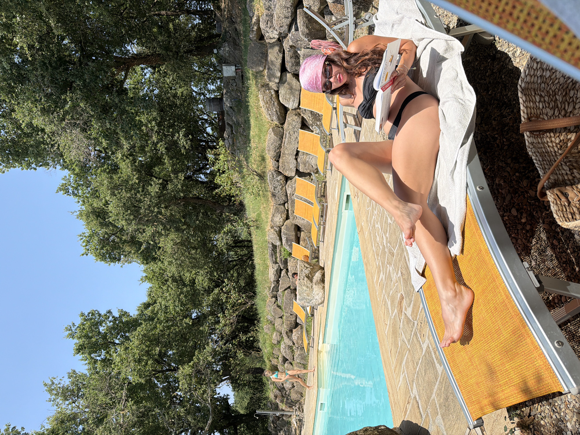 In Sassofortino, Italy, on a sunny day, a woman with long brown hair, wearing a black bikini and a pink bandana, sits on an orange lounge chair by a swimming pool. She is reading a book, holding it open with her left hand and touching the pages with her right hand. Her sunglasses are resting on her nose. She is positioned on a stone patio surrounded by lush green trees and a rock wall. Another woman in a turquoise bikini walks near the edge of the pool in the background. Several yellow lounge chairs are visible near the pool. A woven straw bag rests on the ground near the foreground.
