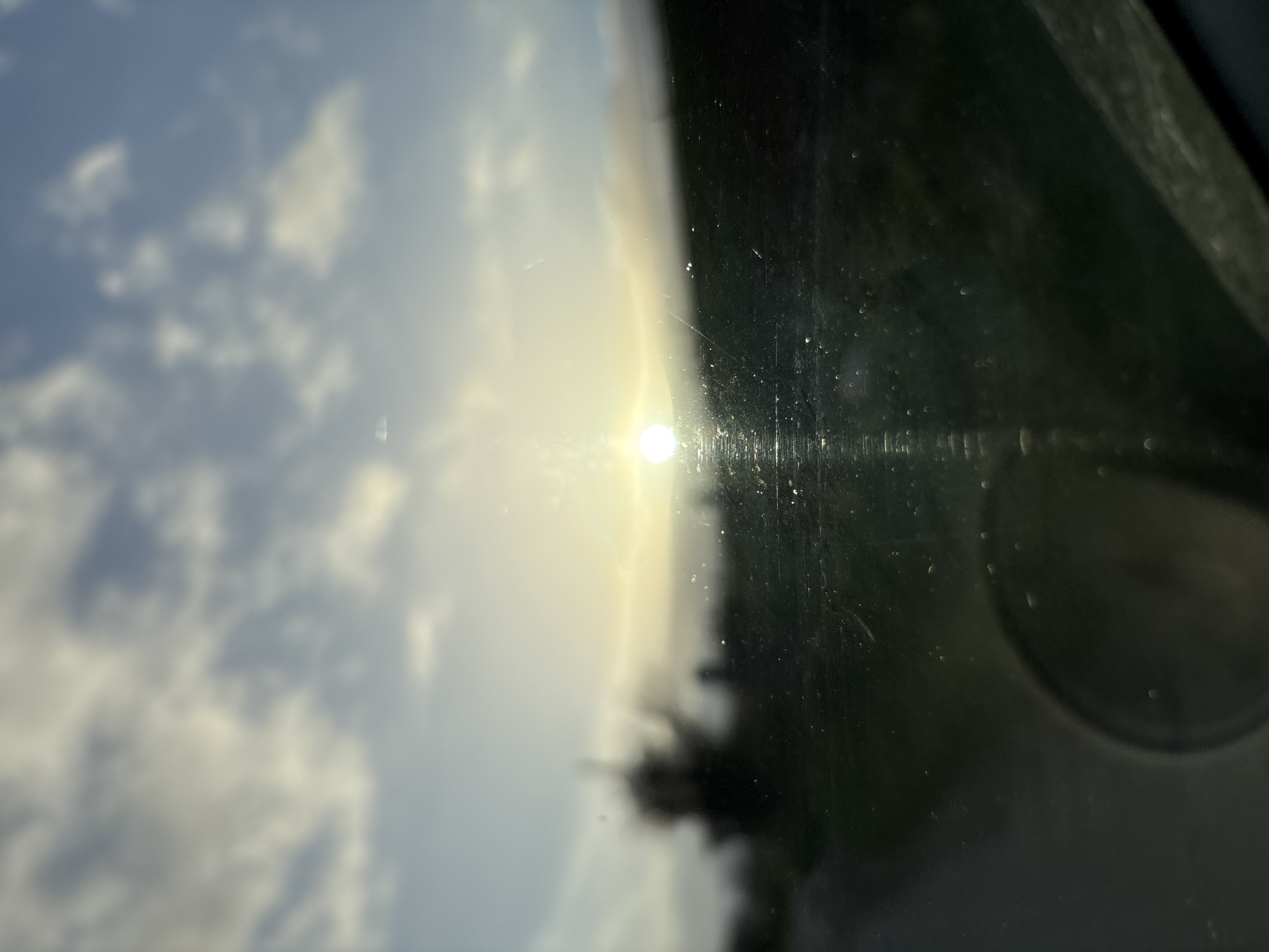 The image depicts an out-of-focus, low-light scene observed through a dirty or scratched glass surface, likely a vehicle windshield, in Shelve, United Kingdom. A bright, circular sun is prominently positioned near the horizon in the left-center, emitting strong glare and vertical light streaks across the viewing surface. A faint halo or lens flare surrounds the sun. The sky above consists of blurry blue with scattered white and grey clouds. The foreground and midground are indistinct, showing a dark, blurry landmass on the right and a suggestion of a road or path leading off to the left, alongside a dark, silhouetted tree-like shape. In the lower-left quadrant, a circular reflection or object, possibly a side mirror, is visible. The bottom edge reveals an indistinct dark structure, likely part of a vehicle's interior. No individuals are present. The environmental conditions suggest either sunrise or sunset.