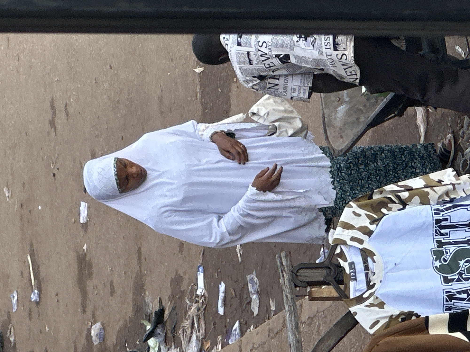 A woman wearing a white hijab and a loose white garment stands in an outdoor marketplace. Her head is bowed, and she is looking down. She carries a white bag over her left arm. To her right, a young person wearing a shirt with a newspaper print walks away from the camera, pushing a wheelbarrow. In the foreground, clothing items are displayed on hangers. The ground is dirt and appears littered with debris and paper. The setting is consistent with the city of Bukuru, Nigeria, where outdoor markets are common.