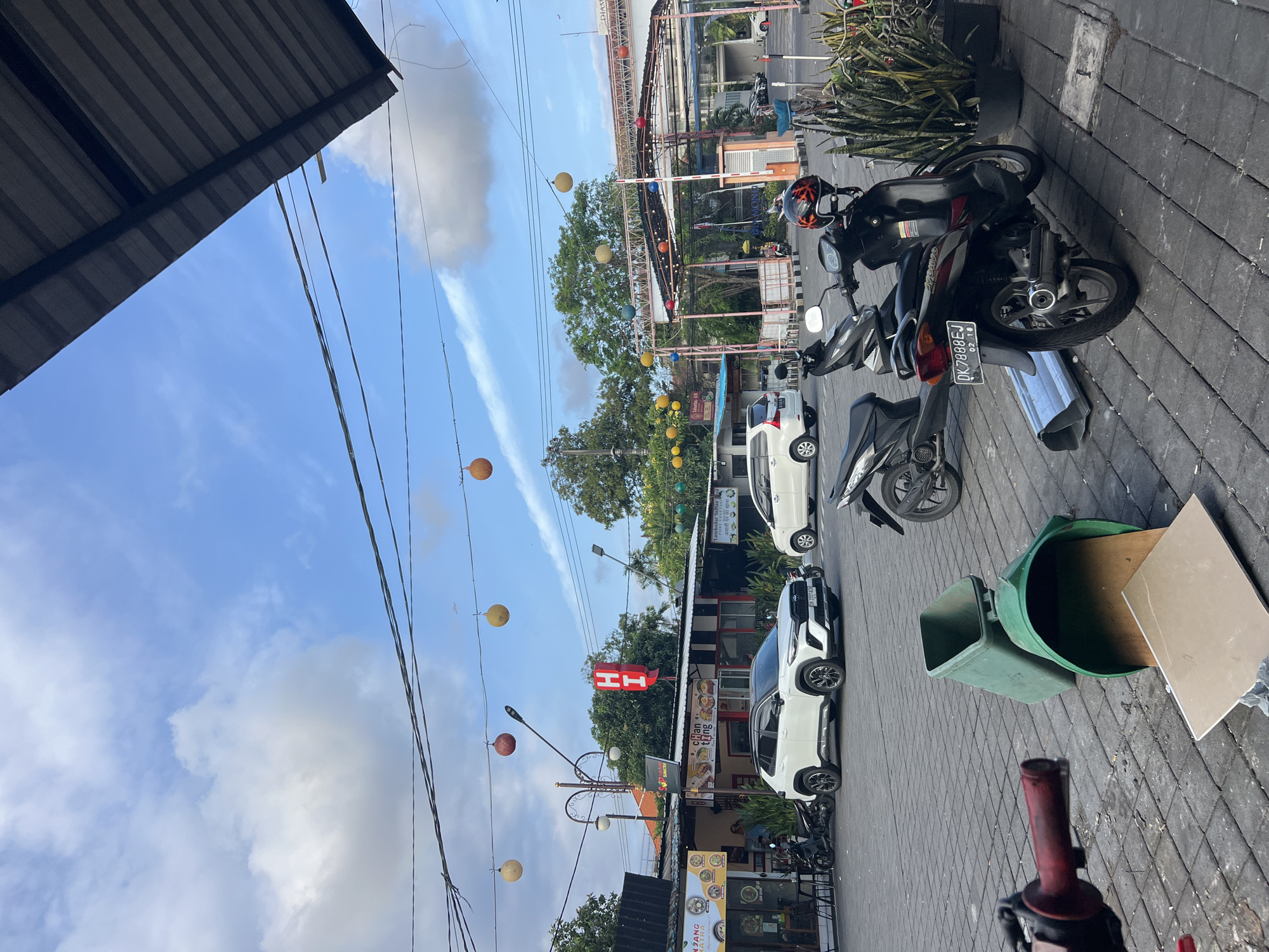The image depicts an outdoor scene in Kuta, Indonesia, likely a commercial or street-side area. A white SUV is parked in the foreground, facing slightly right. To its left, a smaller dark-colored scooter is parked. Behind the white SUV, another white vehicle, possibly a minivan, is visible. Further back, a building with red "HI" lettering and other signage is present, along with trees and a covered structure with colorful lights. Overhead, power lines crisscross the sky, which is partly cloudy blue. On the ground in the lower center of the image, a green trash can is tipped over, spilling its contents, including a piece of what appears to be wood or cardboard. Another scooter is parked in the right foreground, with a helmet on its seat. The ground is paved with dark gray cobblestones.