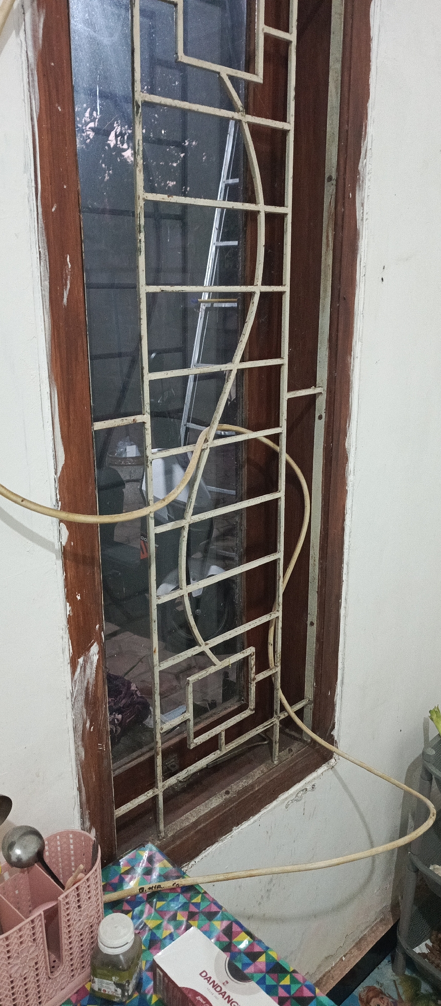 The image shows a close-up view of a window with a white metal grate. The window frame is made of dark wood. Reflections in the glass show trees and what appears to be a ladder inside. A light-colored cable is draped around the grate. In the foreground, on a windowsill, is a pink plastic utensil holder with a ladle, a small glass bottle with a white lid, and a box with the text "DANDANG" visible. The wall to the right of the window is painted white and shows some signs of wear. The scene is likely indoors, possibly in a residential setting in Arjasa, Indonesia, given the context.
