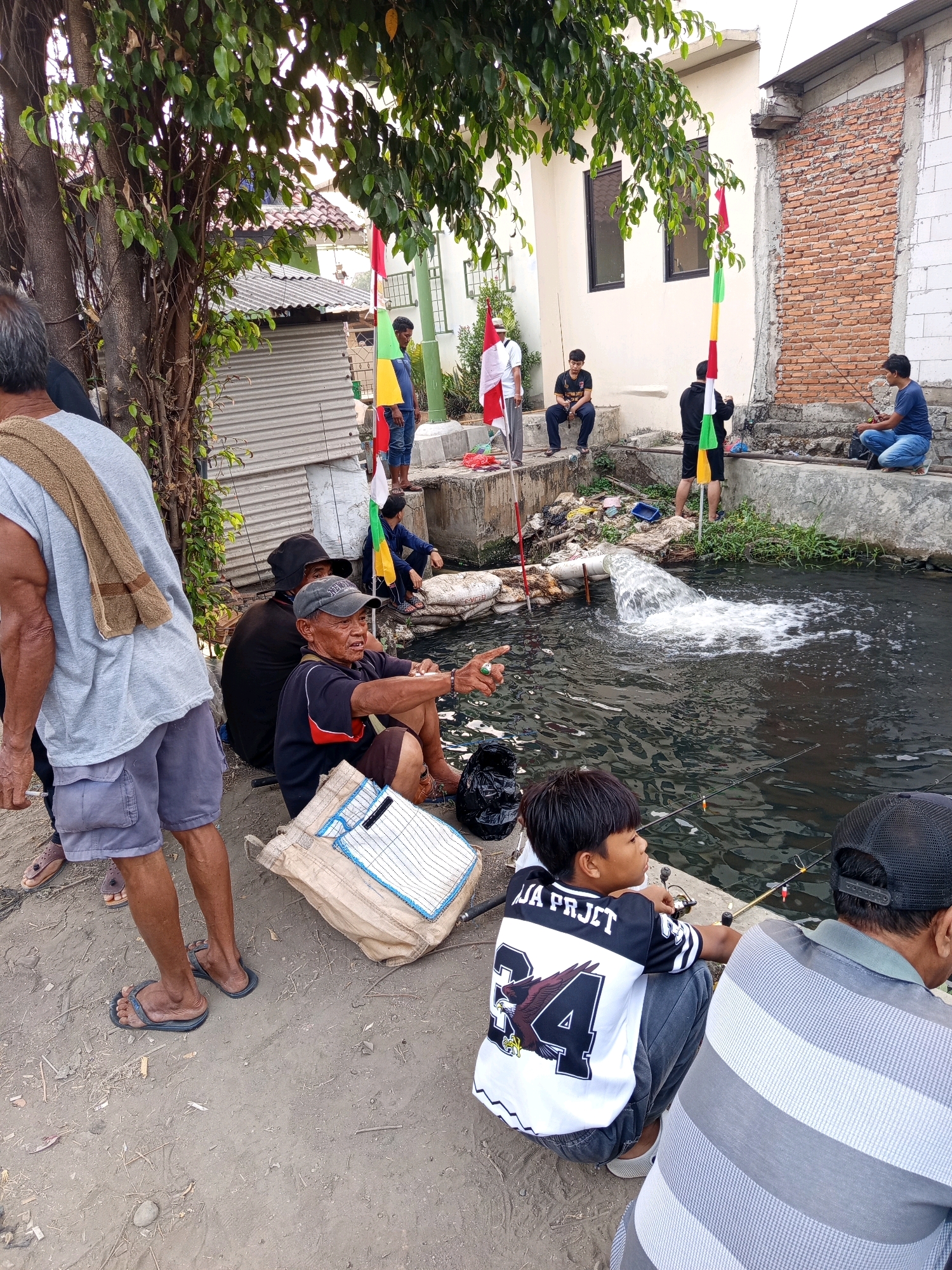 In Karawang, Indonesia, a group of men and a boy are fishing by a murky, flowing canal. The scene is characterized by urban debris, including plastic bags and bottles, scattered along the canal's edge and on the adjacent bank. Red, white, and green flags, likely representing Indonesian national colors, are prominently displayed, tied to poles along the canal.
Several individuals are actively engaged in fishing, with fishing rods extending into the water. One man, seated on the bank, appears to be gesturing towards the canal. Others are positioned on a concrete ledge overlooking the water, some with their fishing gear. A large pipe is visibly discharging water into the canal, creating a significant disturbance on the surface.
The individuals are dressed in casual attire, with some wearing t-shirts and shorts, while others are in longer pants. The ground is a mixture of dirt and gravel. The background shows residential buildings, including one with exposed brickwork and another with a corrugated metal exterior, suggesting an urban or semi-urban environment. The overall impression is of a community gathering for a recreational activity in a somewhat neglected urban setting.