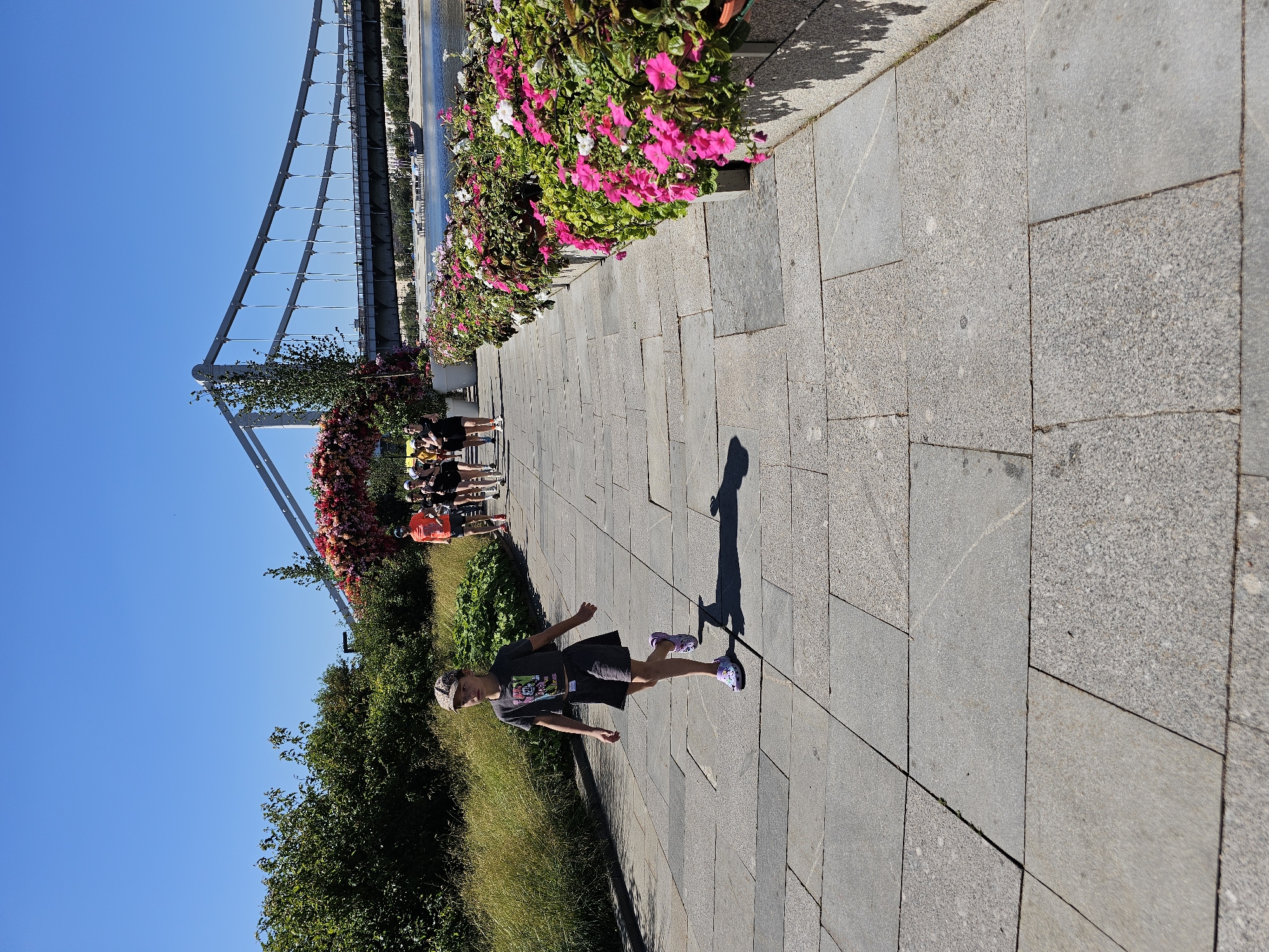 In an outdoor urban setting under a clear blue sky, a young girl wearing a cap, t-shirt, and skirt walks on a paved walkway. She is in the foreground, with her body angled slightly away from the camera. Behind her, a group of people, some wearing orange shirts, are walking in the same direction. The walkway is lined with vibrant pink and white flowers in planters. In the background, a suspension bridge spans a body of water. Lush green trees and foliage are visible along the left side of the walkway and behind the bridge. The scene appears to be a public park or promenade.
