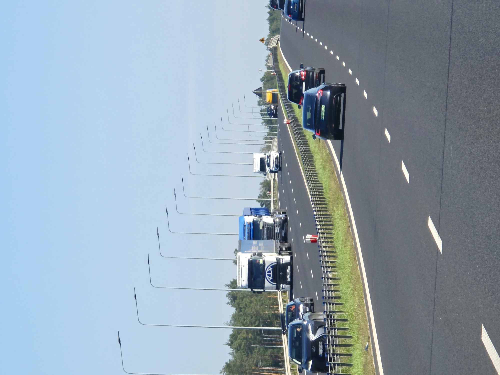 This is a high-angle, eye-level, full shot of a multi-lane highway with a clear blue sky and scattered clouds above. A line of tall streetlights runs along the left side of the highway. The highway has asphalt paving and is divided into multiple lanes by white dashed lines. A metallic guardrail separates the highway from a grassy shoulder. In the foreground, on the right side of the highway, a dark blue sedan is driving. Behind it, a black SUV is visible. Further ahead on the highway, a white truck with a blue trailer is driving in the right lane, followed by a blue dump truck in the left lane. Several other cars and trucks are also visible on the highway in the distance. The surrounding environment is mostly flat with a line of trees visible on the horizon to the left.
