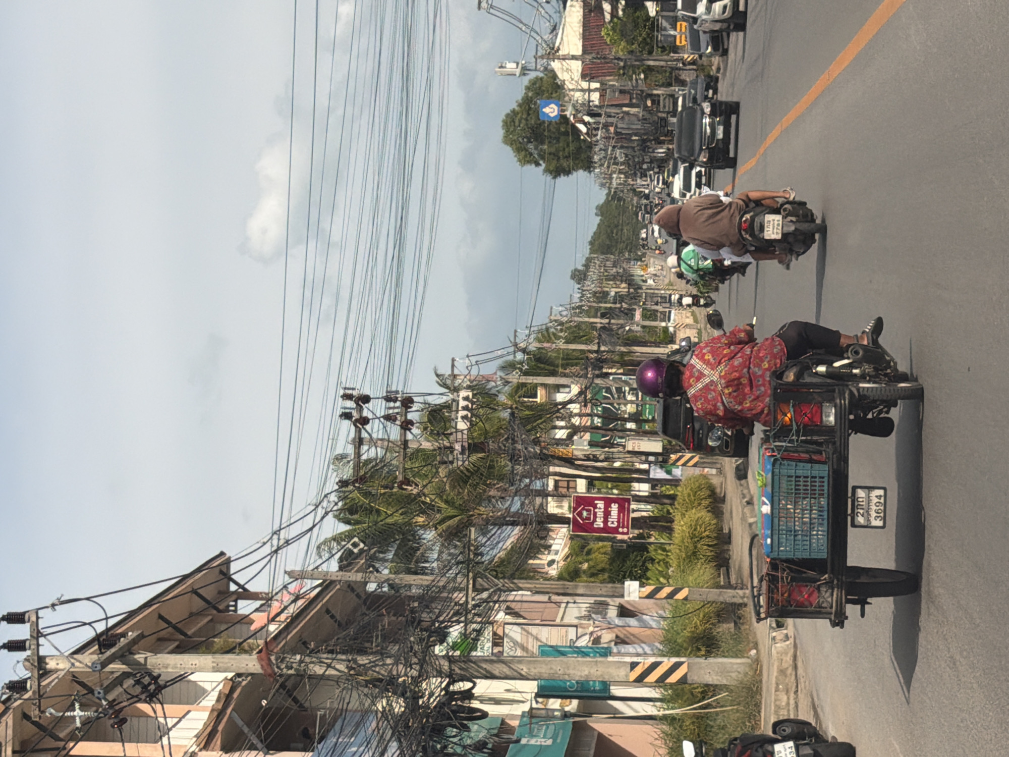 The image depicts a street scene in Baan Bo Phut, Thailand, characterized by a dense network of overhead utility wires and a procession of motorcycles. In the foreground, a motorcycle with a sidecar is prominently displayed, carrying blue plastic crates. The driver, wearing a colorful patterned shirt and a purple helmet, is operating the vehicle. Behind this motorcycle, another motorcycle carrying two individuals is visible, one wearing a brown hooded garment. Further down the road, a variety of vehicles, including cars and more motorcycles, are present, navigating the traffic. The buildings lining the street are typical of a commercial area, with shops and signs, including one for a "Dental Clinic." Palm trees are interspersed with the urban architecture, suggesting a tropical location. The sky is overcast with some clouds. The overall impression is of a busy, everyday street environment in a Thai town.