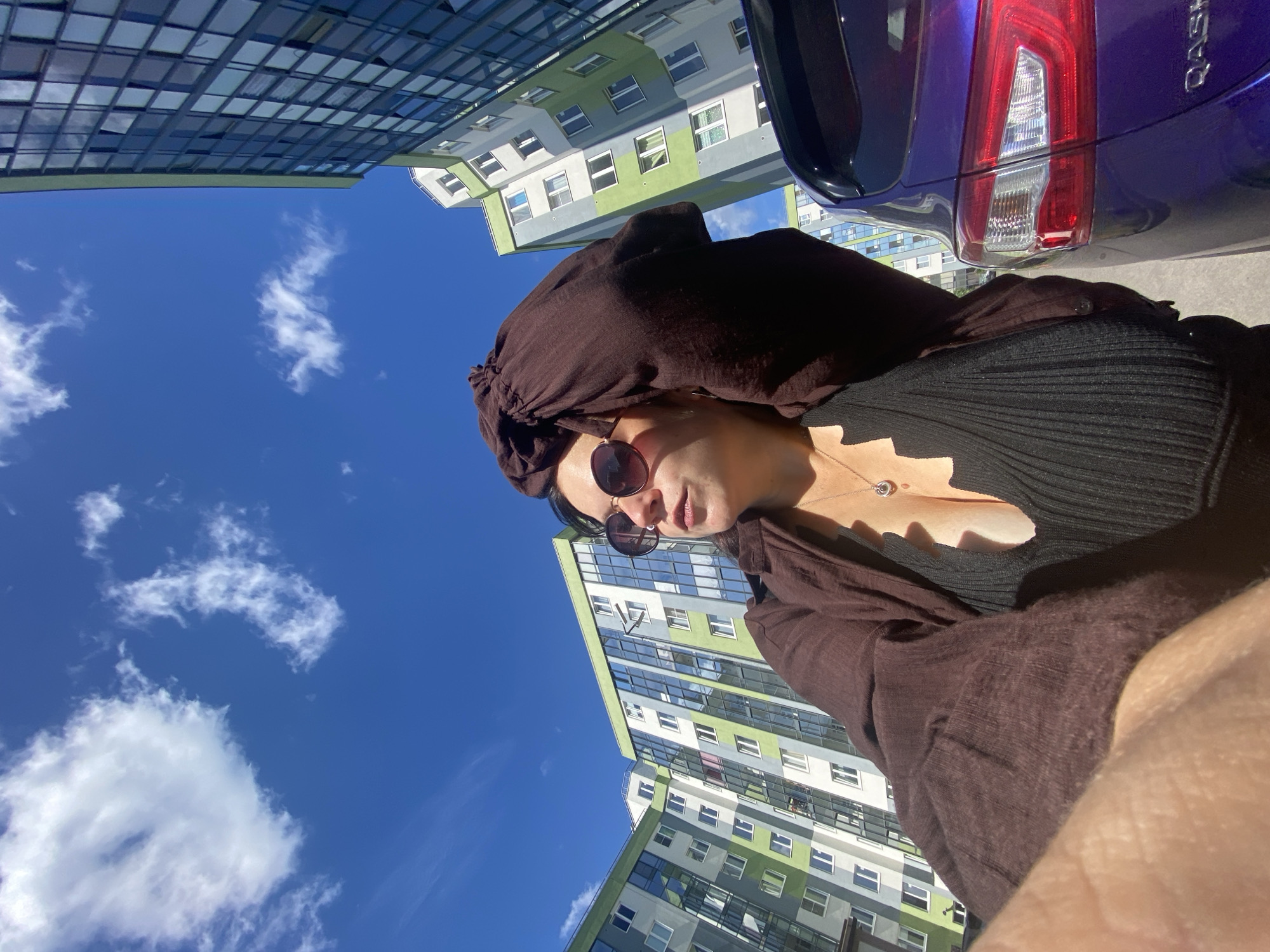 In Yanino-1, Russia, a low-angle shot captures a woman in sunglasses and a brown garment posing in front of modern apartment buildings under a bright blue sky with scattered white clouds. She has her left arm raised, holding her headscarf. A purple car is parked to her right. The buildings feature a mix of glass and colored panels.