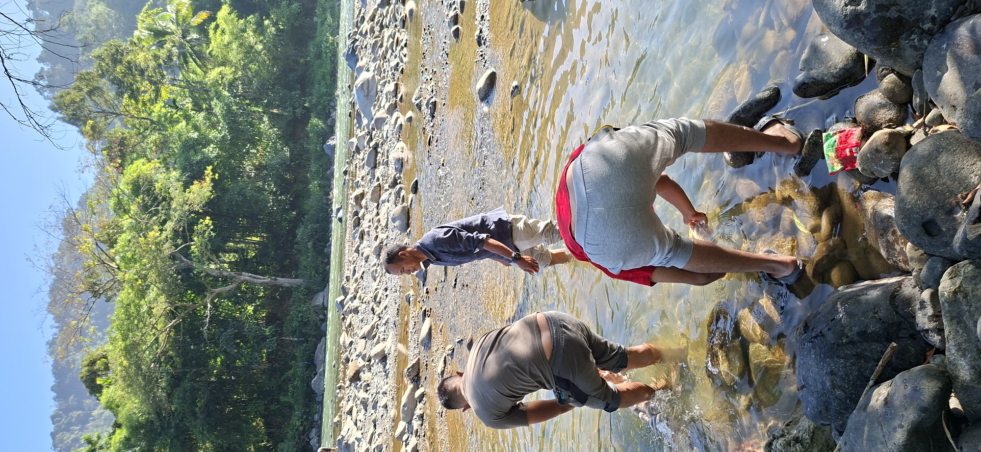 Three men are in a shallow, rocky river in the city of Mane, Indonesia. The man on the left wears a grey t-shirt and grey shorts. The man in the middle wears a dark blue long-sleeved shirt and light-colored pants. The man on the right wears a red t-shirt and grey sweatpants. The man on the right is bent over, seemingly looking at something in the water. The water is clear and reflects the sunlight. The riverbed is covered in smooth, grey stones of various sizes. In the background, a lush green forest covers the hills under a clear blue sky. Several trees are visible at the edge of the forest, and some branches extend into the foreground. There is a small, colorful package lying on the rocks in the foreground.