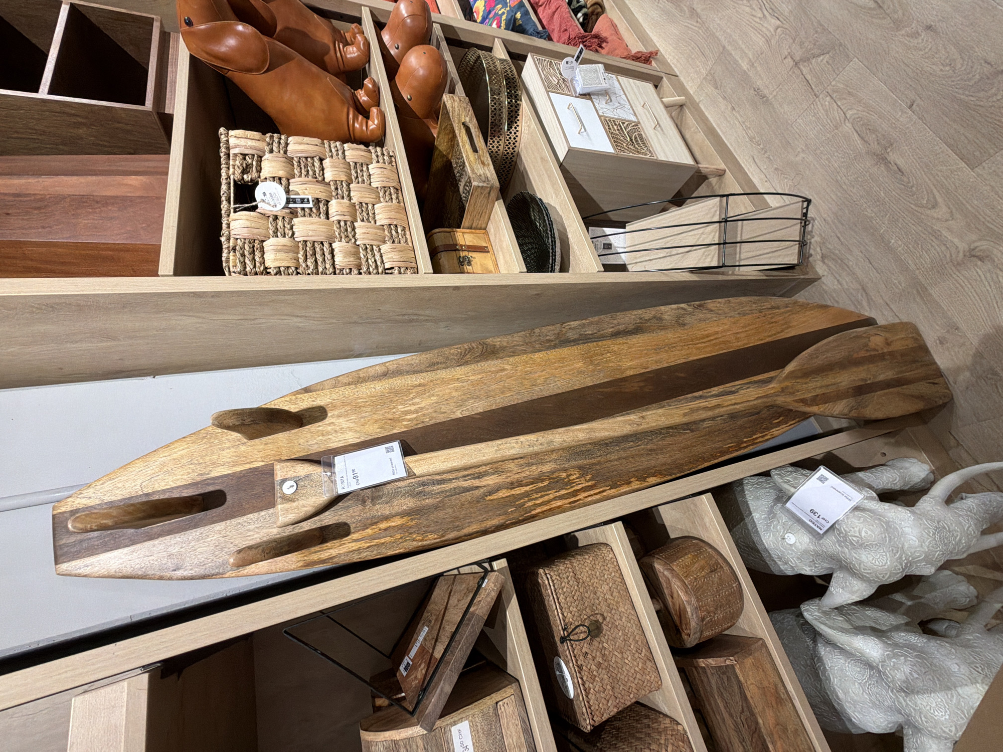 The image shows a retail display of home decor items in a store. In the foreground, a large, rustic wooden surfboard with a paddle rests against a shelving unit. To the right, a tall, narrow shelving unit displays various items including woven baskets, brown leather-like decorative objects resembling a dachshund and perhaps some other animals, and small wooden drawers. A tag on the wooden surfboard reads "ROSITA CHF 91.10". Further down the shelves, there are more baskets, a small wooden chest, a dark green plate, and a decorative brass tray. On the lower shelves, two large white decorative elephant figures are visible, one with a tag that reads "MATEO CHF 139". The flooring appears to be light-colored laminate or wood. The lighting suggests an indoor setting, likely a furniture or home goods store. The background shows more wooden shelving and displays.