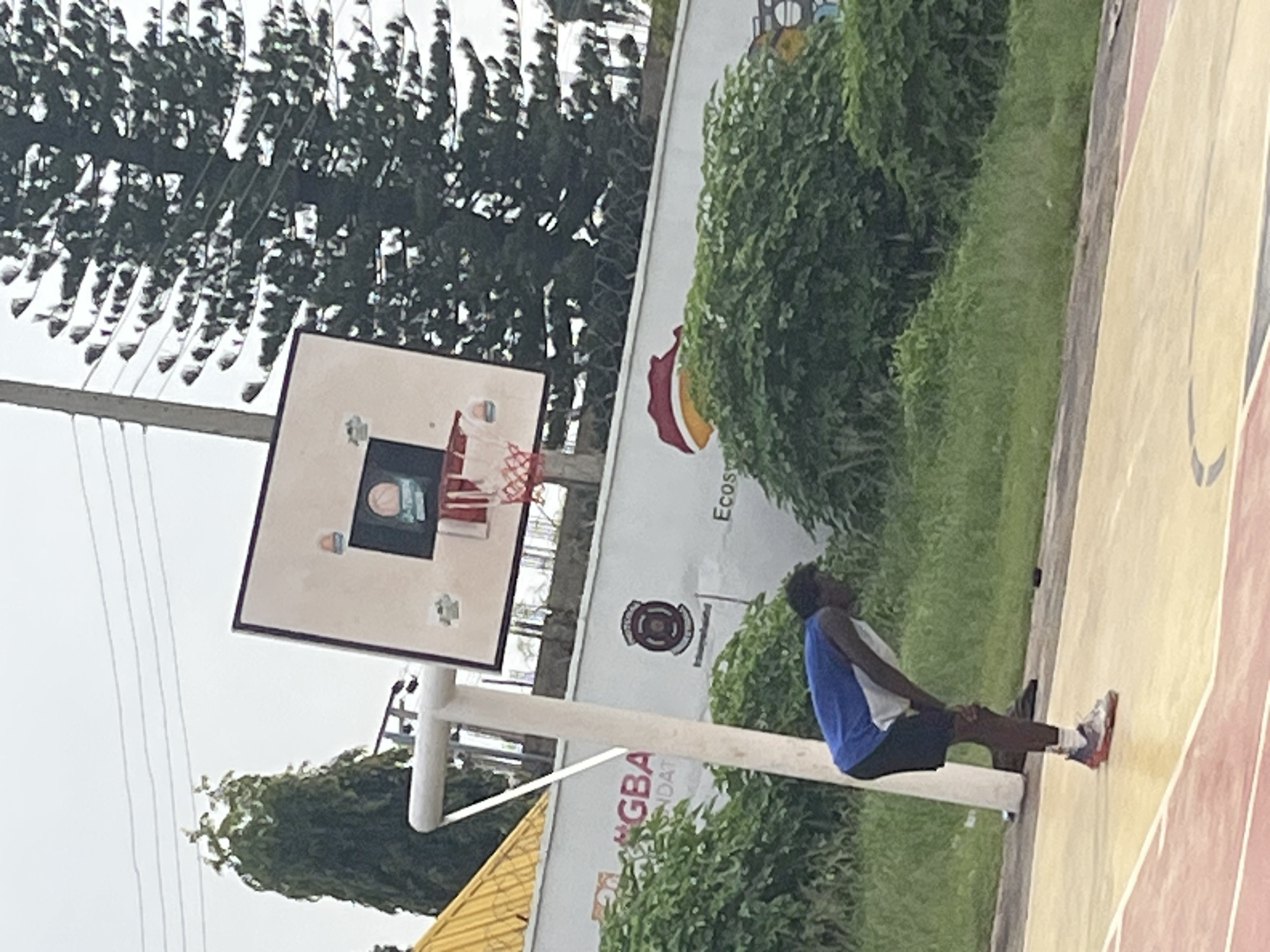 A young Black male wearing a blue and white athletic tank top and black shorts is bent over on a yellow and red outdoor basketball court, with his hands on his knees. He is positioned in the lower left quadrant of the image, with his left foot slightly forward. In the background, a white basketball hoop with a red net stands on a pole. Behind the hoop is a white wall with various logos and text, including "GBA FOUNDATION". Lush green bushes and trees are visible behind the wall, and a large evergreen tree is on the left side of the image. The sky is overcast and appears grey. The location is identified as Gura Top, Nigeria.
