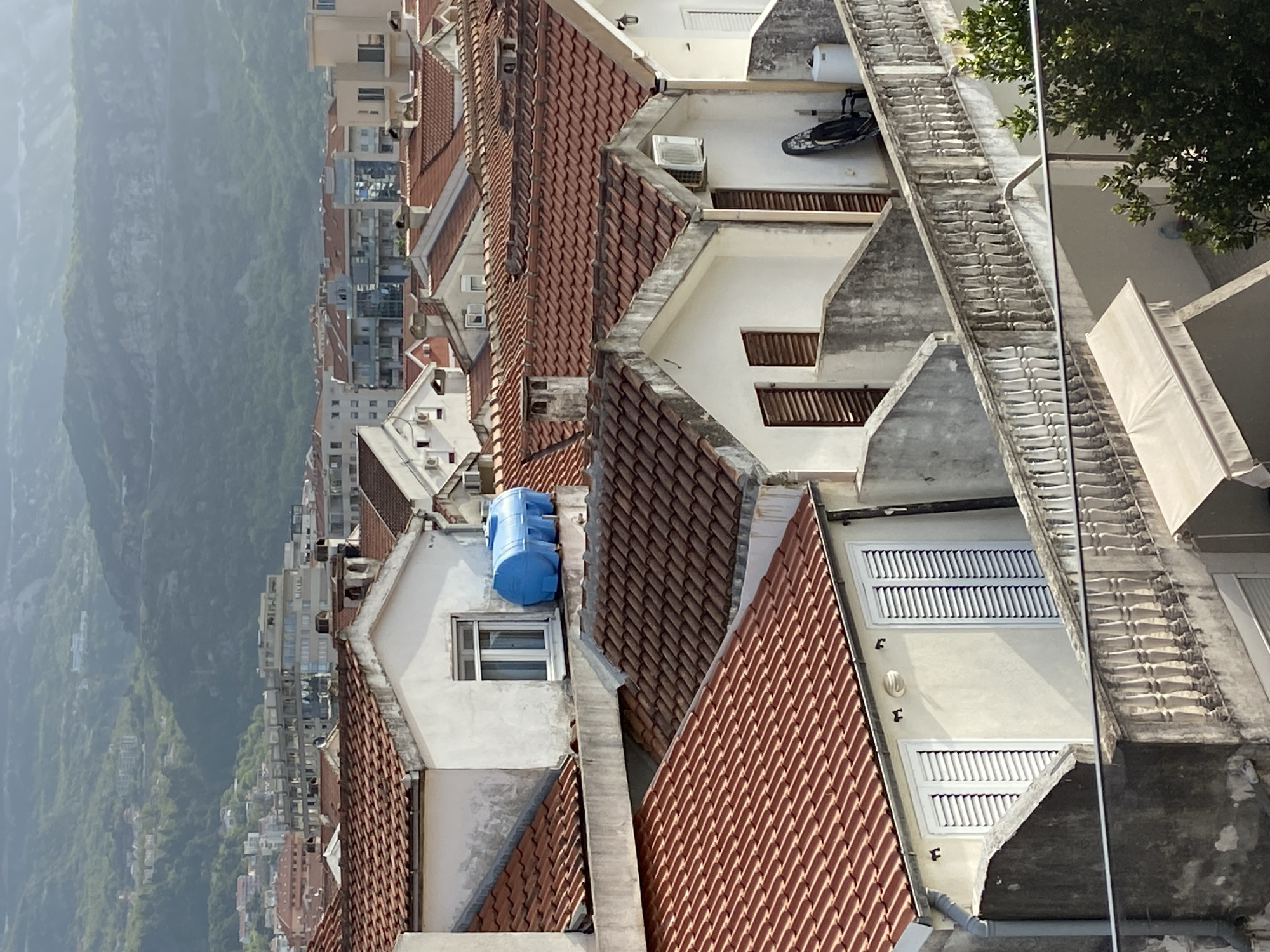 An aerial view shows tightly packed buildings with red tiled roofs in the city of Budva, Montenegro. A mountain range shrouded in a misty haze stretches along the left edge of the frame. The architecture is characterized by white facades and visible air conditioning units. Some buildings feature water tanks and others have shuttered windows. The perspective is slightly askew, and a portion of a balcony with an awning is visible in the lower right corner.