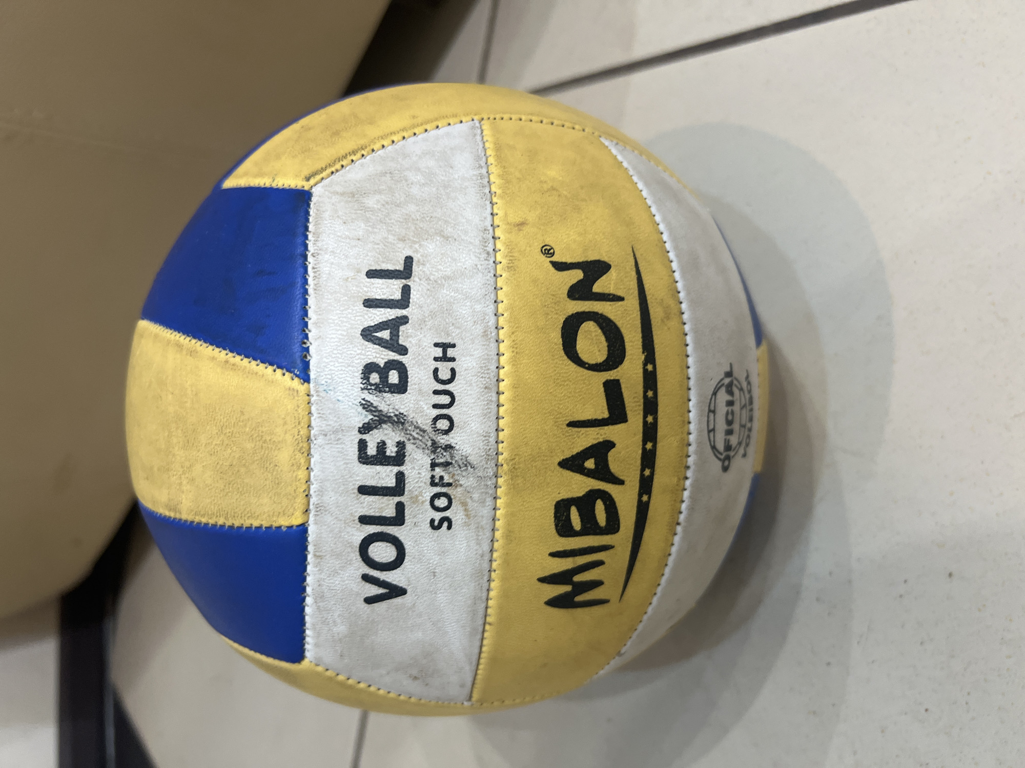 A volleyball, colored yellow and blue, rests on a tiled floor. The ball has the words "VOLLEYBALL SOFT TOUCH" and "MIBALON" printed on it. A small logo that appears to read "OFICIAL VOLEIBOL" is also visible. The lighting suggests an indoor setting, possibly a home or sports facility. The image does not provide any information about the location in the city of Трещиха, Russia, as there are no identifiable landmarks or contextual clues present.