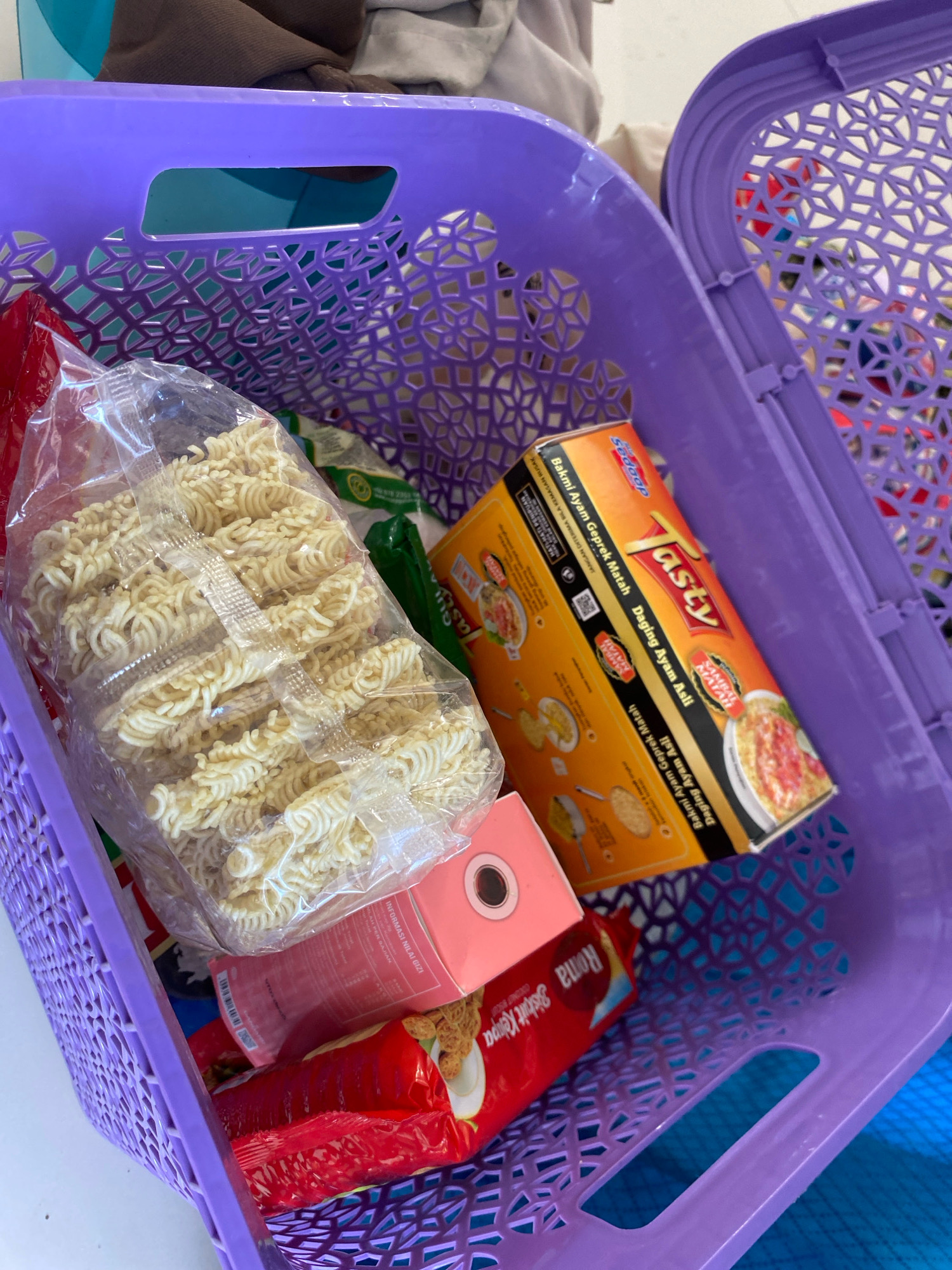 The image shows a close-up, high-angle shot of a purple plastic basket filled with various food items. The basket is positioned on a light blue surface, possibly a table or countertop. Dominating the foreground is a large, clear plastic bag filled with uncooked instant noodles, their coiled strands neatly packed. To the right of the noodles, a rectangular orange box of "Tasty" brand instant noodles, specifically "Daging Ayam Asli" (Authentic Chicken Meat) flavor, is visible. The box has colorful graphics and text. Partially obscured beneath the noodles and orange box is a pink rectangular box with a circular cutout, and below that, a red package of "Roma Biskuit Kelapa" (Coconut Biscuits). In the background, out of focus, are other items of clothing or fabric, suggesting this might be a domestic setting. The overall impression is of groceries or snacks prepared for storage or transport. The image was taken in Gisik Cemandi, Indonesia, but no specific landmarks or environmental features of the city are discernible in this close-up view.