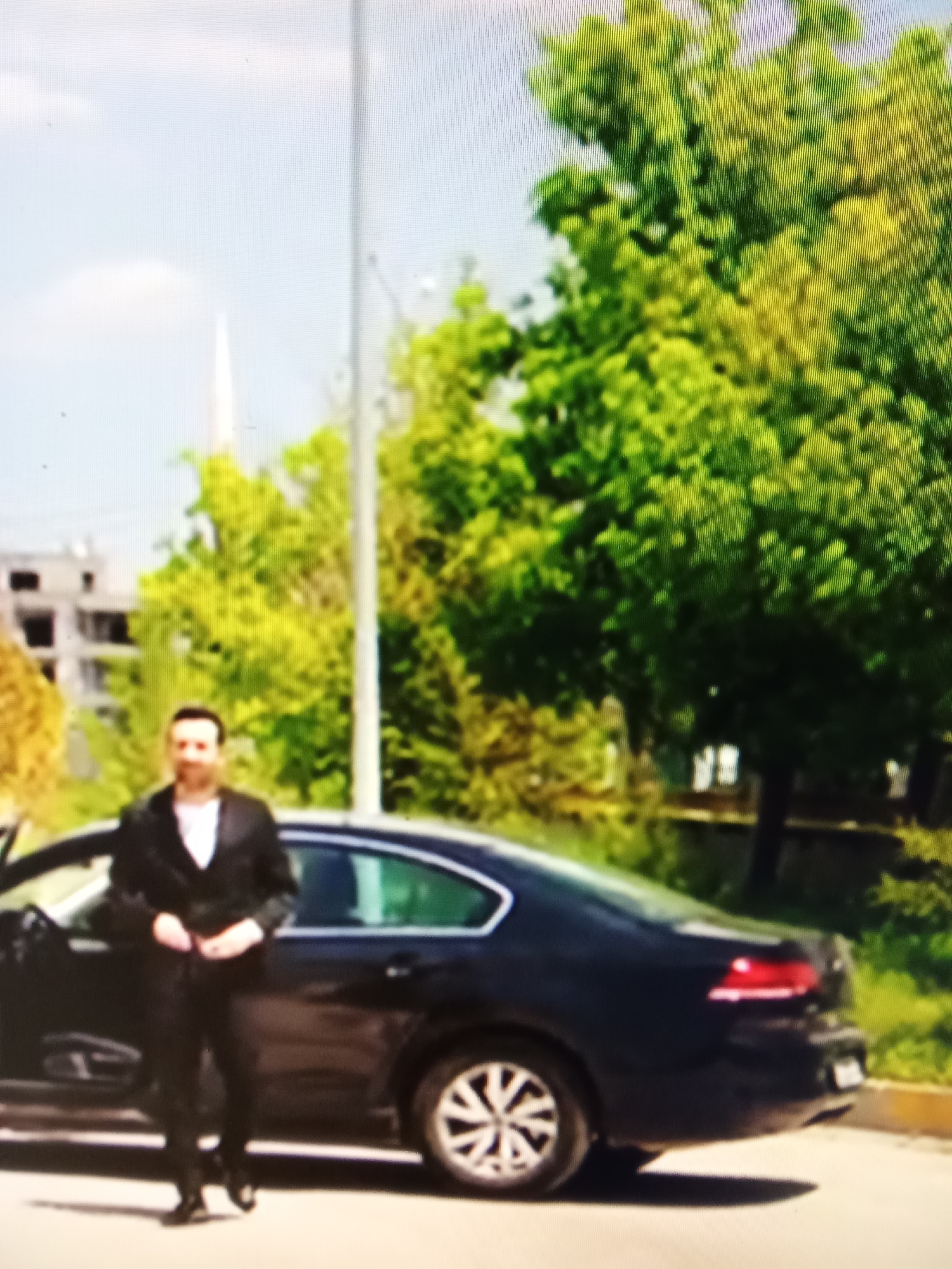 The image shows a man in a suit standing next to a black car. The man is facing the camera and appears to be adjusting his clothing. The car has its driver's side door open. In the background, there are trees and a building that appears to be under construction. A white spire, possibly belonging to a mosque or church, is visible in the distance. The scene is set outdoors on a sunny day. The resolution of the image is low, making it difficult to discern fine details. There is no specific information to determine the exact location within Рощин ауылдық округі, Kazakhstan.