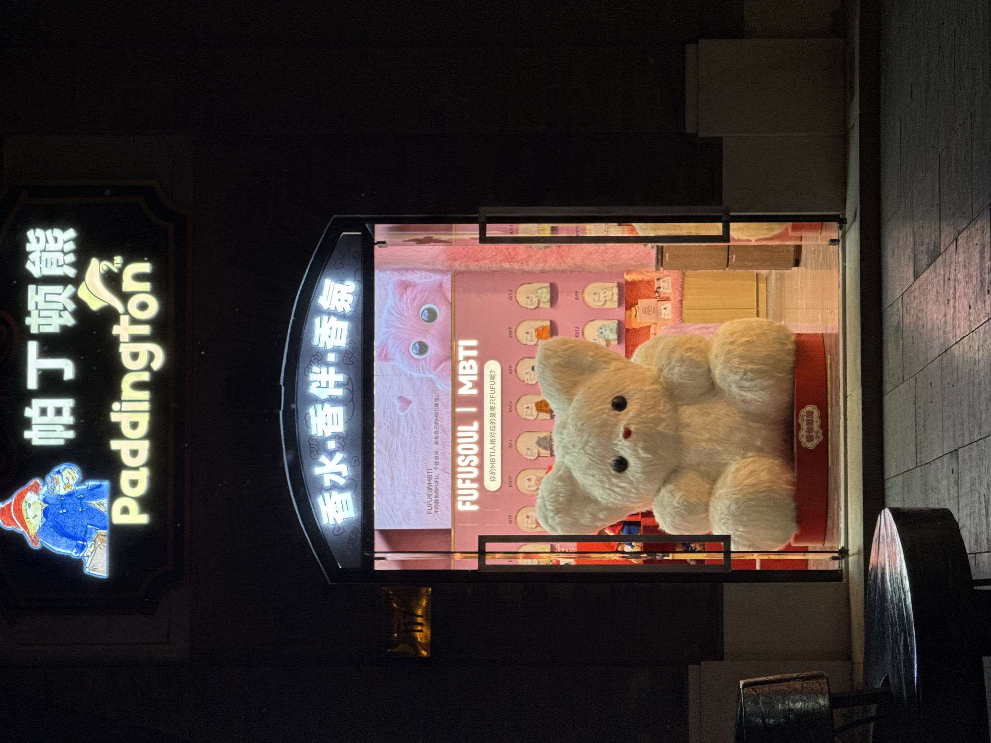 The image depicts a storefront at night in Dongcheng District, China. The storefront is identified by two neon signs: "Paddington" with an image of the character and "FUFO" with Chinese characters. The store window displays a large white plush bear and a pink promotional display featuring the "FUFUSOUL" brand and personality types in the MBTI (Myers-Briggs Type Indicator) format. A pink cat image is included in the background of the display.