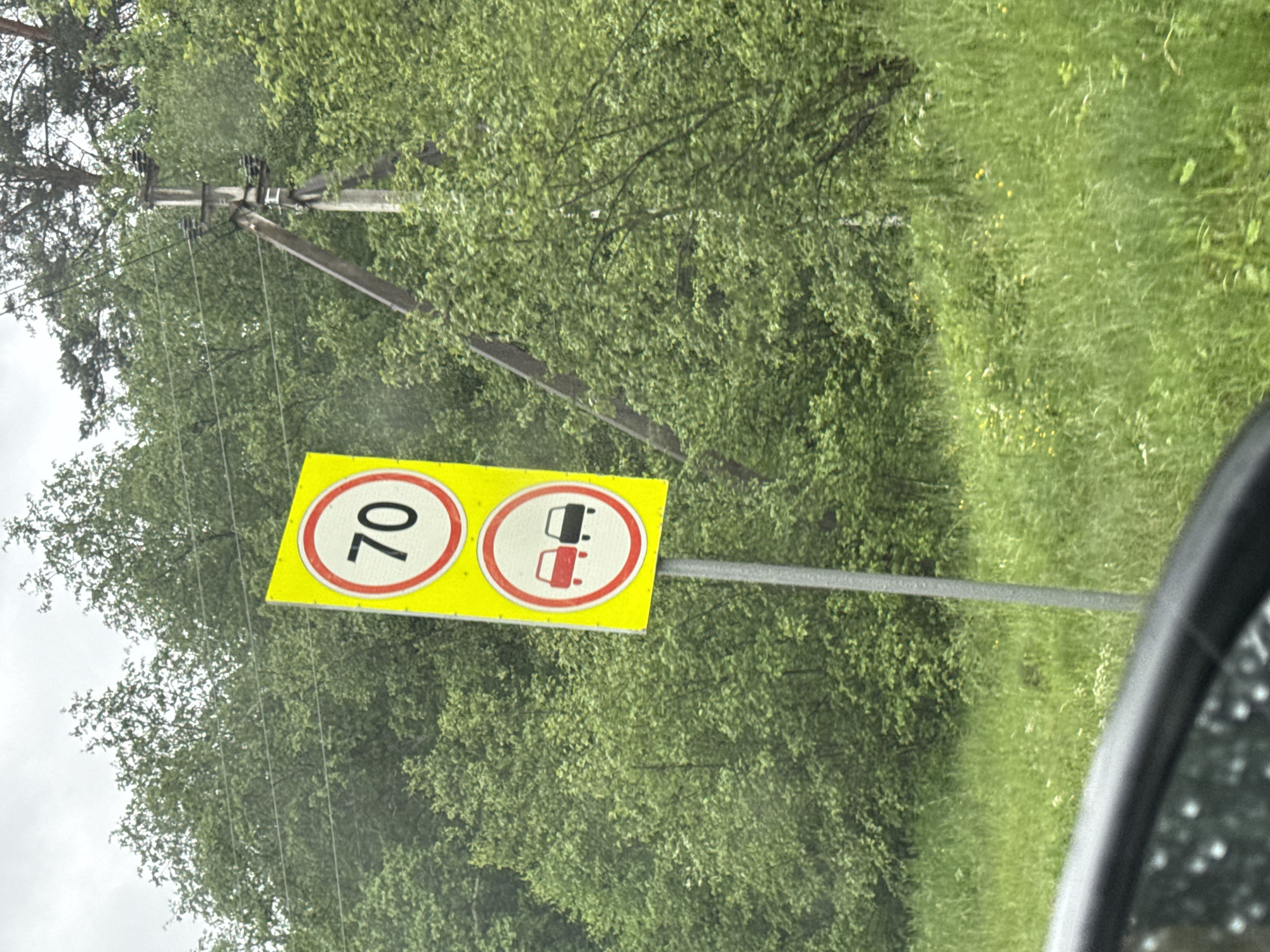 The image displays a roadside scene with two traffic signs on a metal pole, set against a backdrop of dense green foliage and trees. The signs are yellow and rectangular, with the upper sign featuring a red circle and the number "70" in black, indicating a speed limit of 70. Below it, the second sign shows a red circle with a diagonal red bar over two car icons, one red and one black, signifying a prohibition of overtaking. A power line pole with insulators is visible to the right of the signs, and the foreground shows the edge of a vehicle, likely a car, with a wet surface reflecting light. The environment is lush and green, suggesting a rural or suburban setting, possibly during or after rain given the wet appearance of the car's surface. The location is identified as Usmangali, Russia.