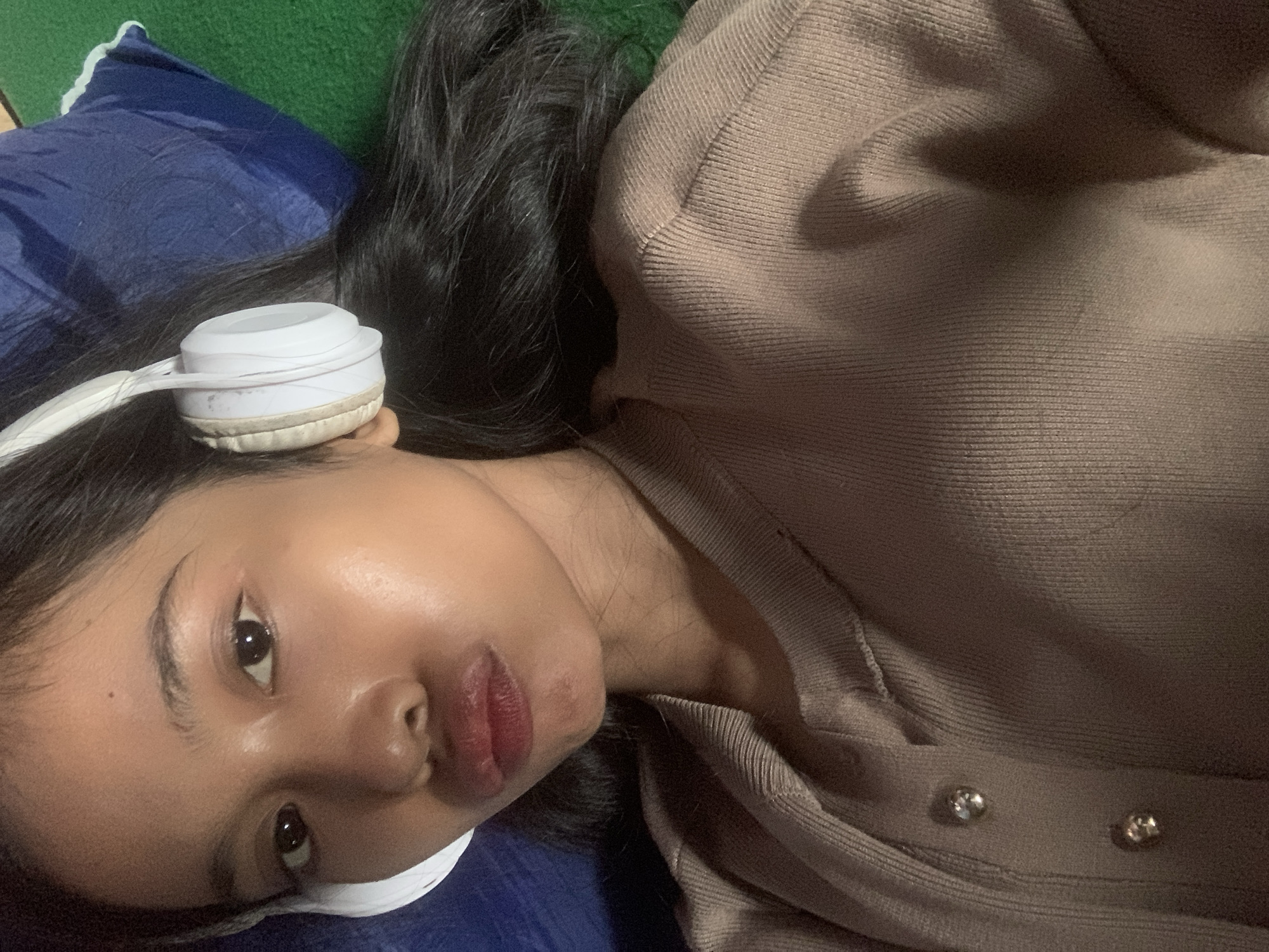 A young woman with dark hair and headphones is lying down, likely in a bedroom in Sungai Meriam, Indonesia. Her head rests on a dark blue pillow, which is on a green surface. She is wearing a light brown sweater with two buttons. The lighting suggests it is nighttime or indoors with artificial light.