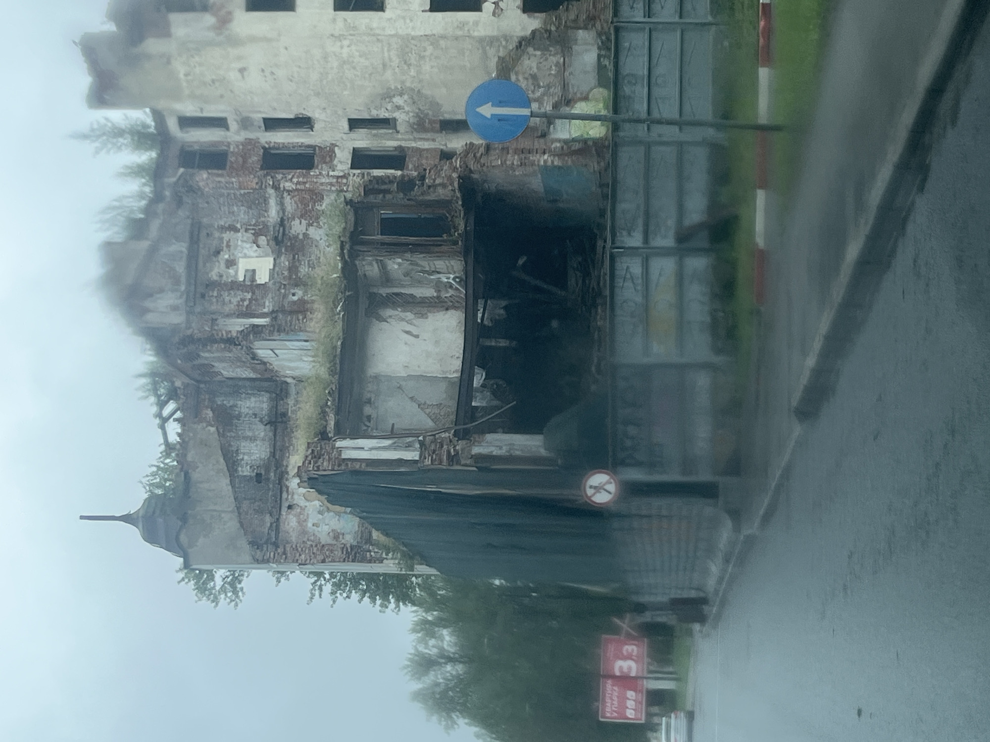 The image shows a severely damaged multi-story building, likely an apartment building, in Saint Petersburg, Russia. The building is in a state of ruin, with its facade crumbling, revealing exposed brickwork and structural damage. Some sections appear to have collapsed, creating large openings. Overgrown vegetation can be seen on the rooftops and within the damaged structure. A dark, tarp-like material is draped over a section of the building, possibly for temporary protection or containment.
In the foreground, a wet asphalt road runs alongside the building, indicating it may have rained recently or is raining. A curb separates the road from a grassy area with a metal fence. A road sign with a white arrow pointing upwards on a blue circular background is visible. Further down the road, red and white striped markers are present. To the left, a white car is partially visible, and behind it, a red billboard with Russian text and the number "3,3" is displayed. A small circular sign with a red border and a diagonal line through a human figure is also visible on the fence. The sky is overcast, contributing to a somber atmosphere. The overall scene depicts an urban environment affected by significant structural decay and possibly past conflict or natural disaster.