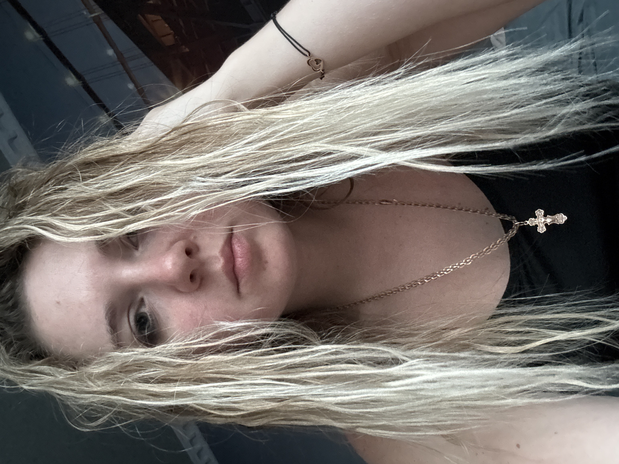 This is a close-up, slightly overhead, sideways portrait of a young woman. She has very long, blonde, somewhat tousled hair that partially obscures her face. Her head is tilted, and her gaze is directed towards the viewer. She is wearing a dark garment, possibly a tank top, and a rose gold chain necklace with a cross pendant. On her left arm, she wears a thin black string bracelet with a small rose gold heart charm.
The background is dark and appears to be indoors, with some blurred shapes suggesting furniture or architectural elements. The lighting is soft, with no strong shadows, indicating it might be daytime or well-lit indoors. The woman's expression is neutral, perhaps a little melancholic or introspective. There is no visible text in the image. The overall mood is intimate and personal.