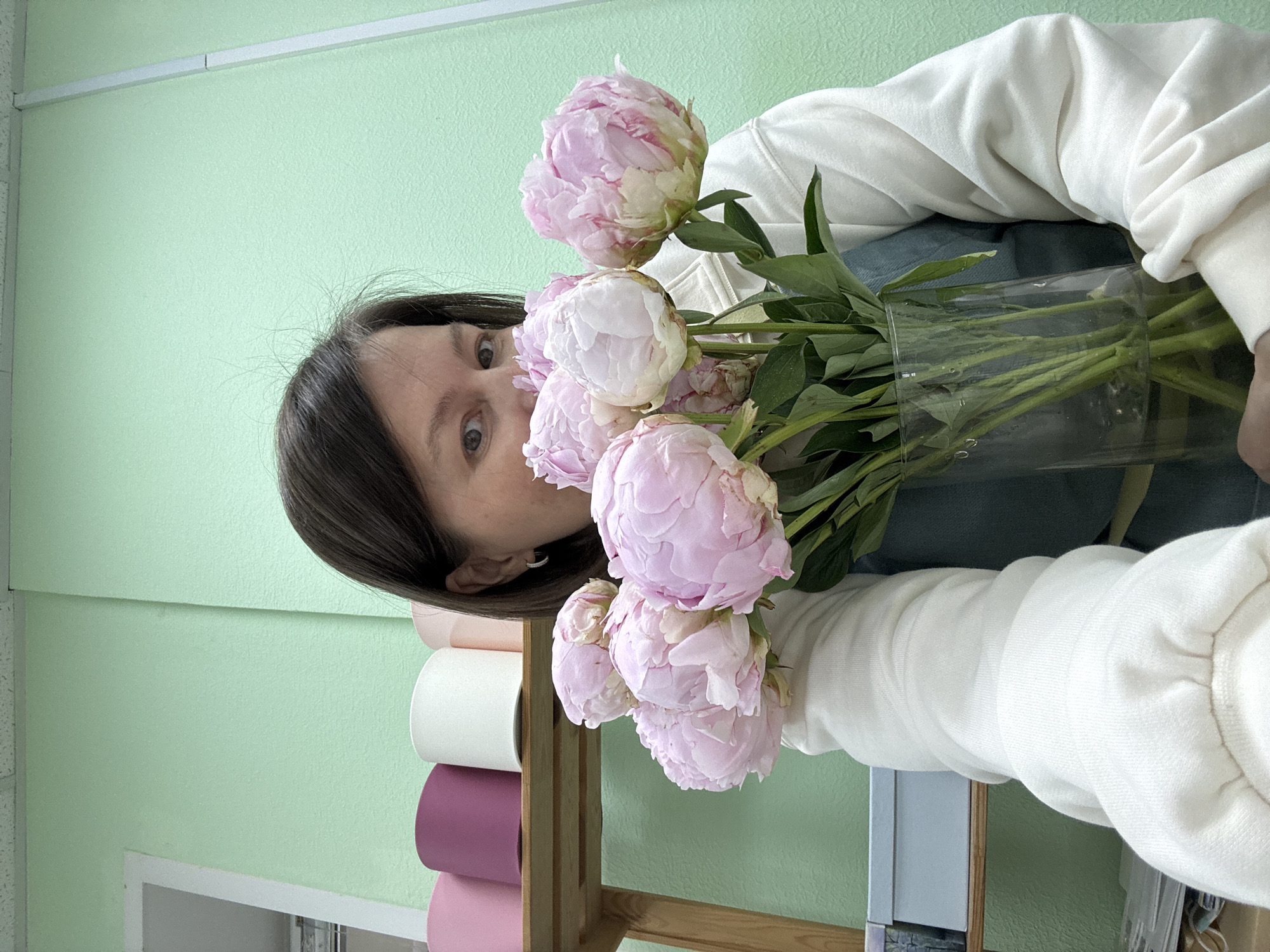 A woman with dark hair is holding a vase of pink peonies. She is wearing a white sweatshirt. Behind her is a light green wall, a wooden rack with rolls of white, pink, and purple paper, and a window. The location is Izhevsk, Russia.