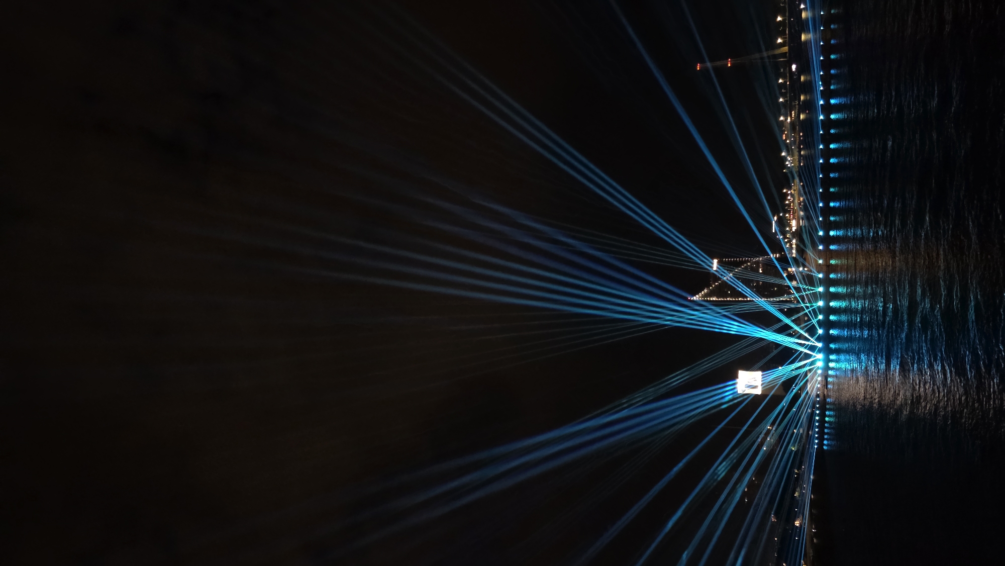 The image captures a nighttime cityscape, with a focus on a dramatic laser light show over the Daugava River in Riga, Latvia. Numerous bright blue laser beams emanate from the riverfront, fanning out upwards and across the dark sky, creating a radial pattern. These beams illuminate the night sky and reflect on the rippling water's surface, creating shimmering green and blue patterns.
In the background, the silhouette of the city skyline is visible. Two prominent structures stand out: a modern skyscraper with a distinctive angular design and a tall, slender tower with a red light at its peak, likely part of a bridge or communication structure. Along the riverfront, a line of lights, possibly streetlights or decorative illuminations, creates a glowing border between the land and the water. A large, square screen or projection is visible in the middle distance, displaying a white image or light. The scene is devoid of any visible people or vehicles, emphasizing the visual spectacle of the light show itself. The overall atmosphere is one of a vibrant, urban nighttime display.