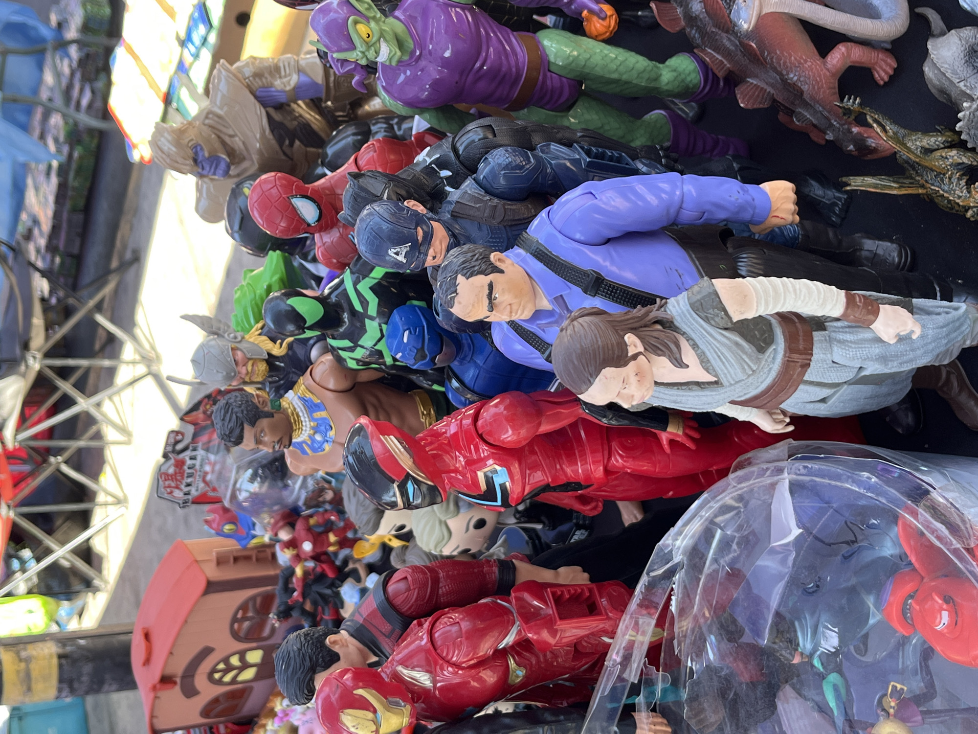 The image shows a collection of various action figures piled together, likely for sale. The figures include characters such as Spider-Man, Captain America, Green Goblin, Star Wars characters, and Iron Man. The setting appears to be an outdoor market stall or table due to the presence of what seems to be a tent structure in the background. The precise location within the city of Unknown, Unknown is not discernible from the visual data.