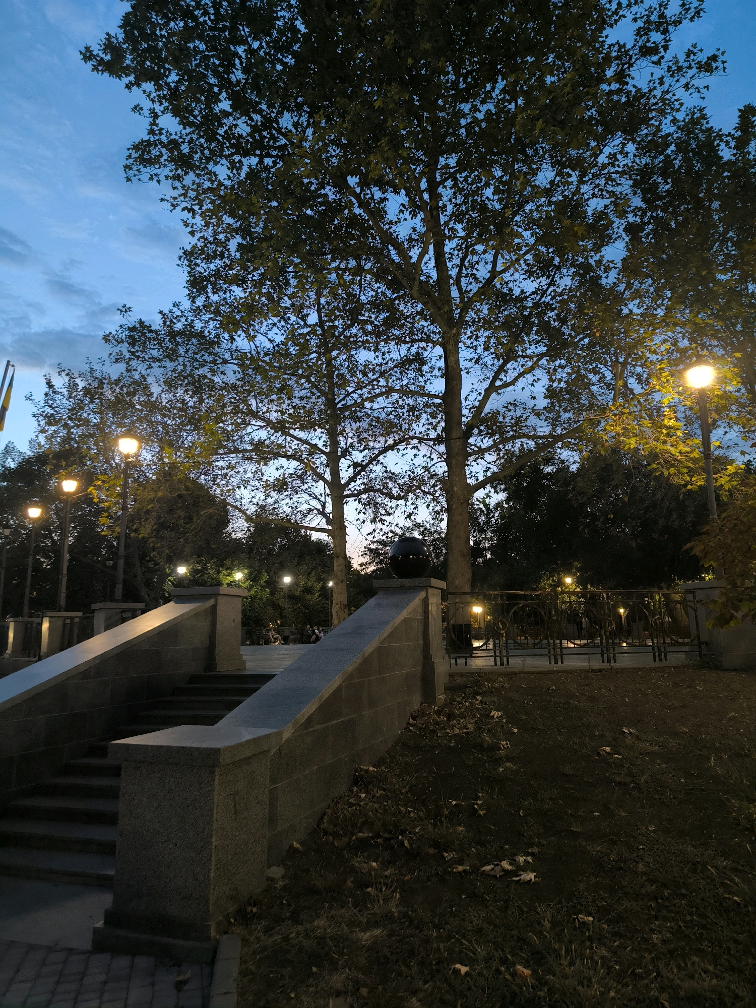 The image depicts an outdoor scene at dusk, likely in a park or public square in Mykolaiv, Ukraine. In the foreground, a stone staircase with granite balustrades ascends to the right, bordered by a paved walkway. Beyond the staircase, trees with sparse foliage are visible, interspersed with ornate lampposts casting a warm yellow glow. A decorative metal fence with intricate scrollwork separates the paved area from a grassy incline dotted with fallen leaves. In the background, the twilight sky is a deep blue with faint cloud formations. A few individuals are faintly discernible in the middle ground, seated or standing near the lampposts. The overall impression is one of a tranquil urban evening.
