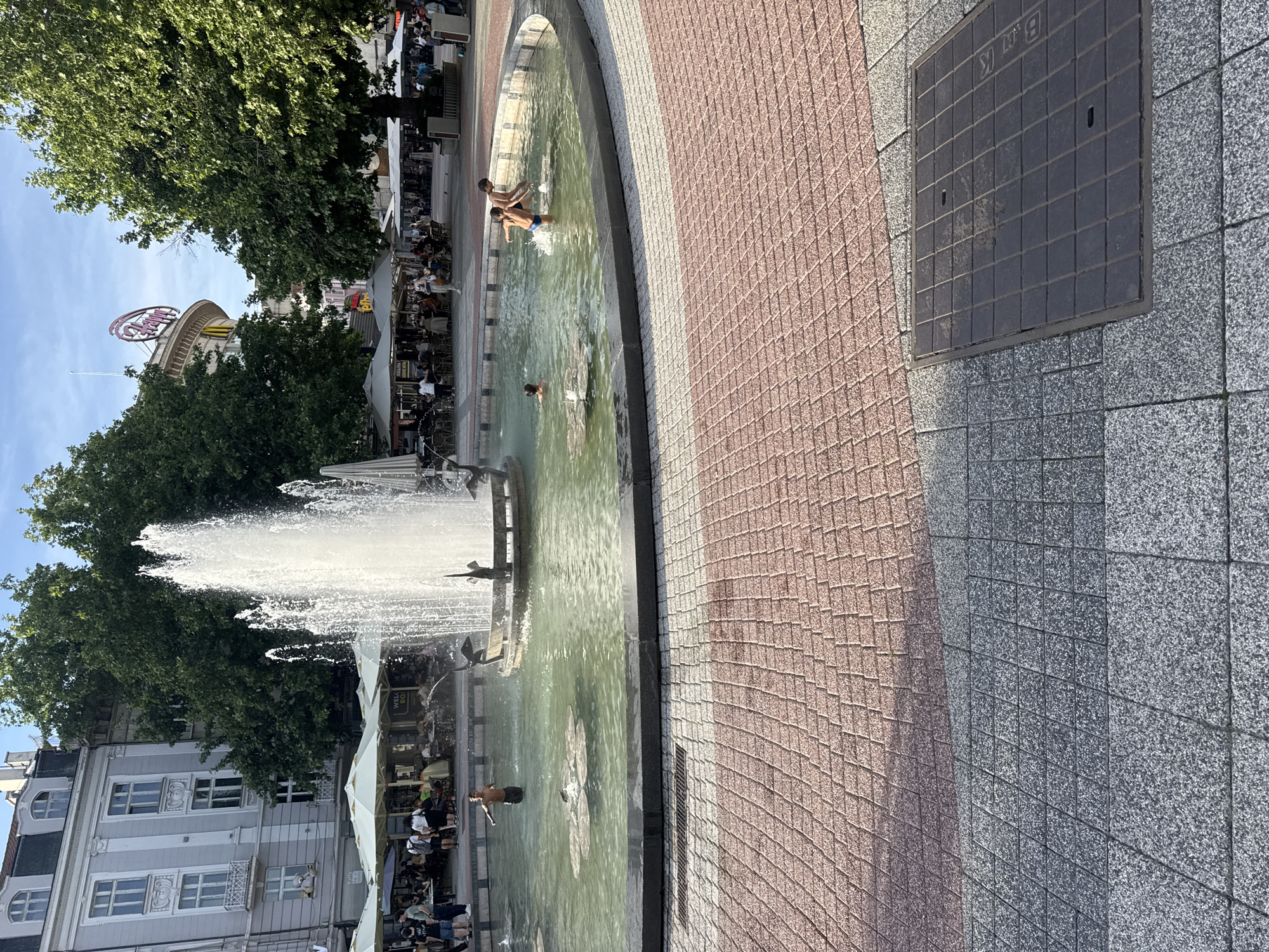 The image shows a circular fountain in Plovdiv, Bulgaria, with several people swimming in it. The fountain is located in a public square surrounded by buildings and trees. There are cafes and restaurants with outdoor seating on one side of the fountain. In the background, the illuminated sign of the Trimontium Princess Hotel is visible. The ground around the fountain is paved with reddish and grey tiles. A square manhole cover is located on the right side of the image.