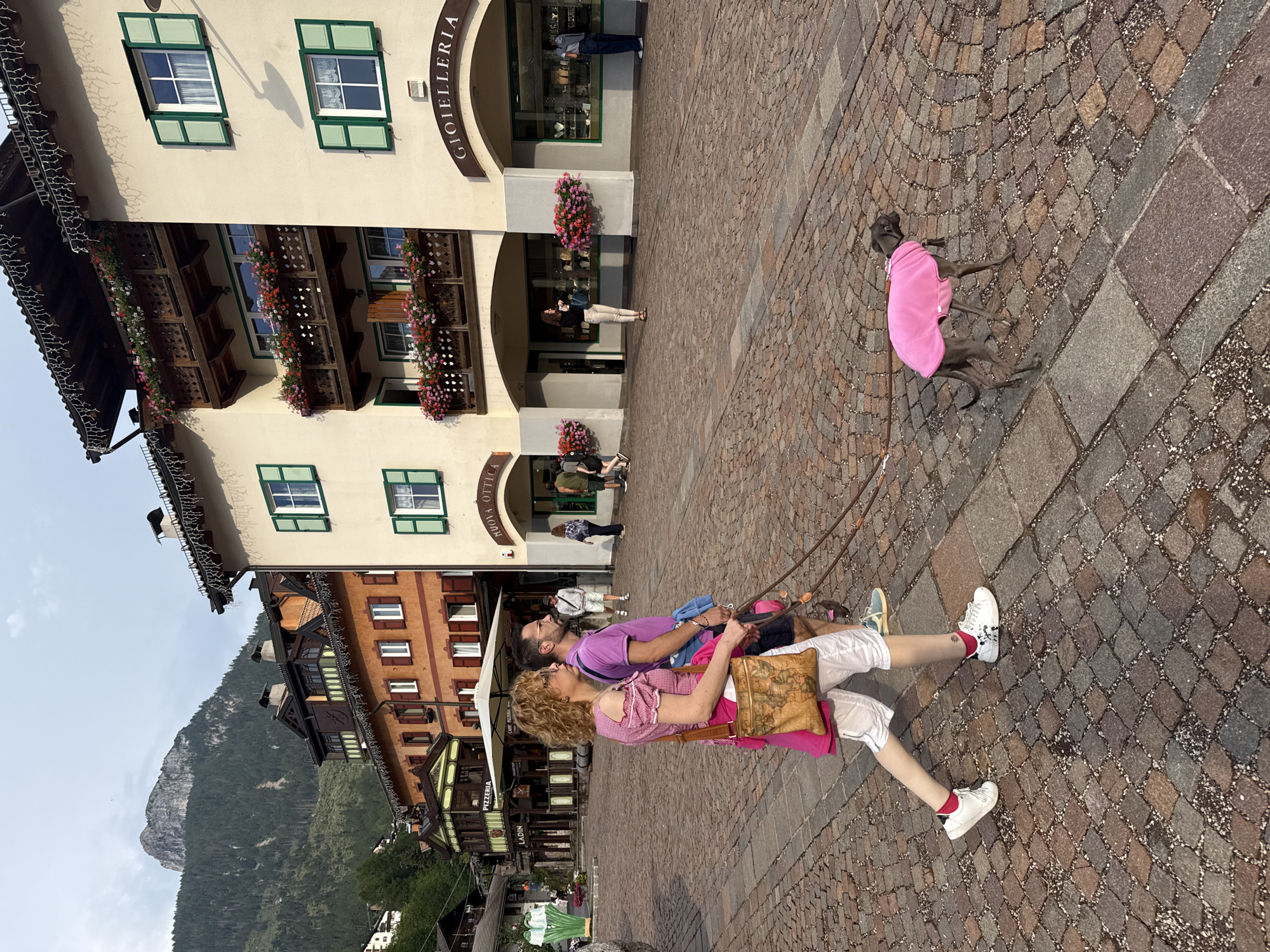 In Moena, Italy, on a cobbled stone street, a couple walks a small grey dog wearing a bright pink coat. The woman, on the left, has curly blonde hair and wears a pink and white checkered shirt, white capri pants, and white sneakers. She carries a tan crossbody bag with a map design. The man, to her right, has dark hair and glasses, and wears a purple polo shirt, dark shorts, and blue sneakers. He holds a dog leash. The dog is an Italian Greyhound. Behind them, a multi-story building with a light-colored facade features balconies adorned with colorful flowers. Green shutters are visible on some windows. The street has a warm, sunlit cobblestone surface. In the background, other people are walking and shopping near storefronts. A prominent mountain peak covered in dense green trees rises majestically in the far distance under a hazy sky. The architecture suggests a European alpine town.