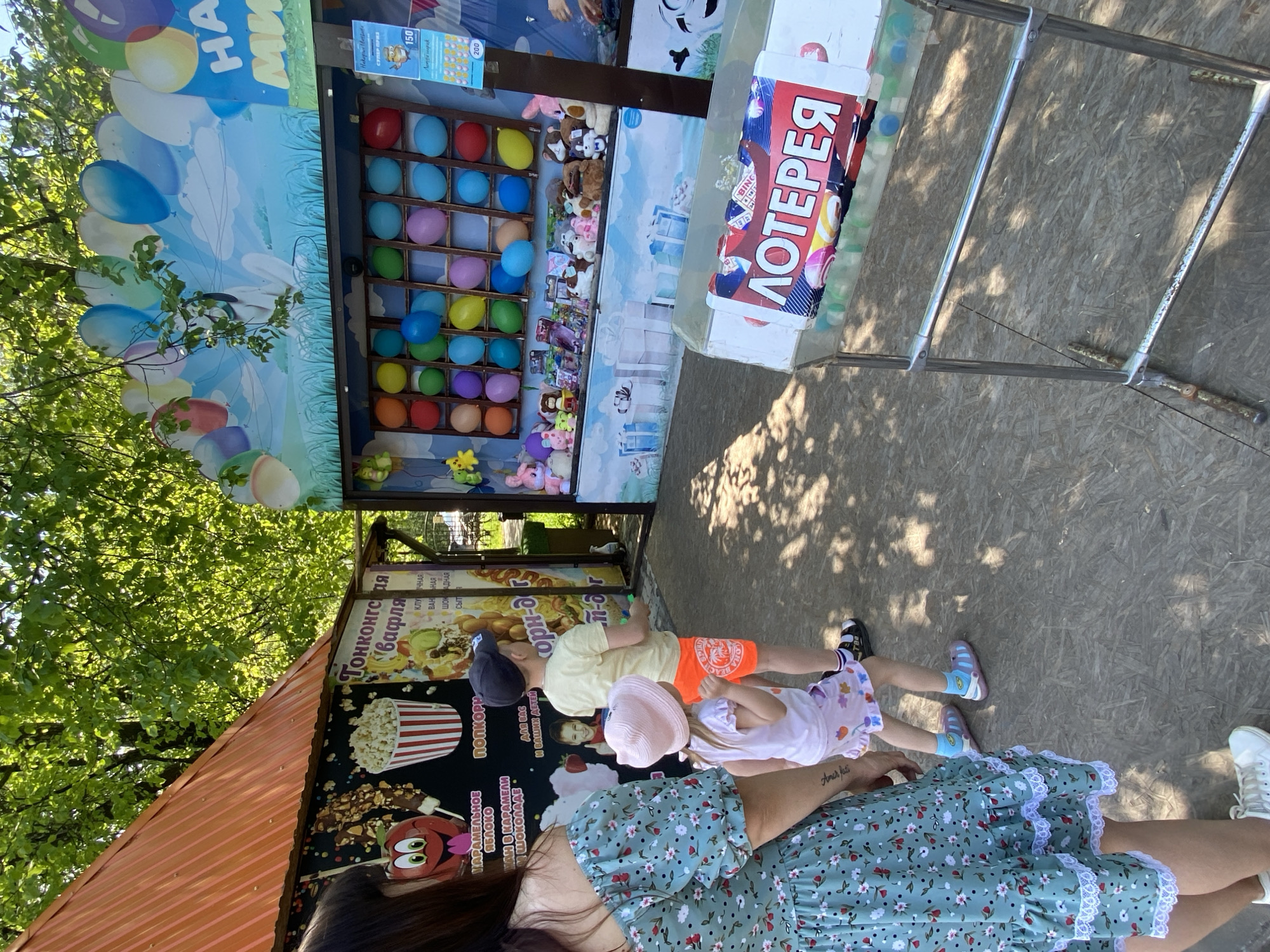 The image captures a scene at an outdoor event or amusement park in what appears to be the city of Chaikovsky, Russia. A woman wearing a green floral dress stands near two children, one in orange shorts and a yellow shirt, and the other in a pink hat and a light-colored dress. They are positioned near a game stand that features balloon popping and prizes. The stand is decorated with a colorful balloon graphic and has a sign labeled "ЛОТЕРЕЯ" (Lottery). Additional stands in the vicinity include a food vendor offering "ГОНКОНГСтая вафля" (Hong Kong-style waffle) and other treats. The environment includes natural elements such as trees, casting shadows on the scene.