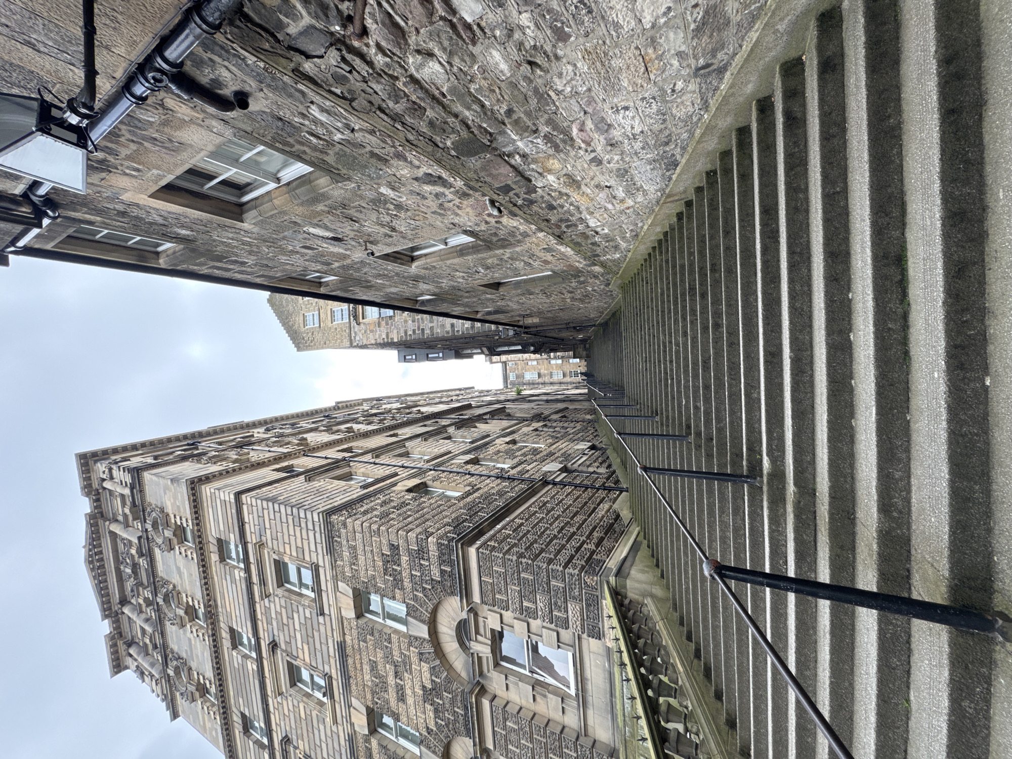 The image shows a long, wide, outdoor stone staircase ascending between two imposing stone buildings in the city of Edinburgh, United Kingdom. The staircase is made of large, gray concrete slabs and is flanked by black metal handrails attached to black metal posts. The building on the left is tall and ornate, with multiple stories featuring many windows and decorative architectural elements. The building on the right is also constructed of stone, appearing older and more rugged, with visible mortar between the stones and a prominent downpipe. The sky is overcast and a pale gray. In the distance, further staircases and buildings can be seen, suggesting a densely built urban environment with significant elevation changes. The perspective is from the bottom of the stairs looking upwards. No people are visible in the frame.