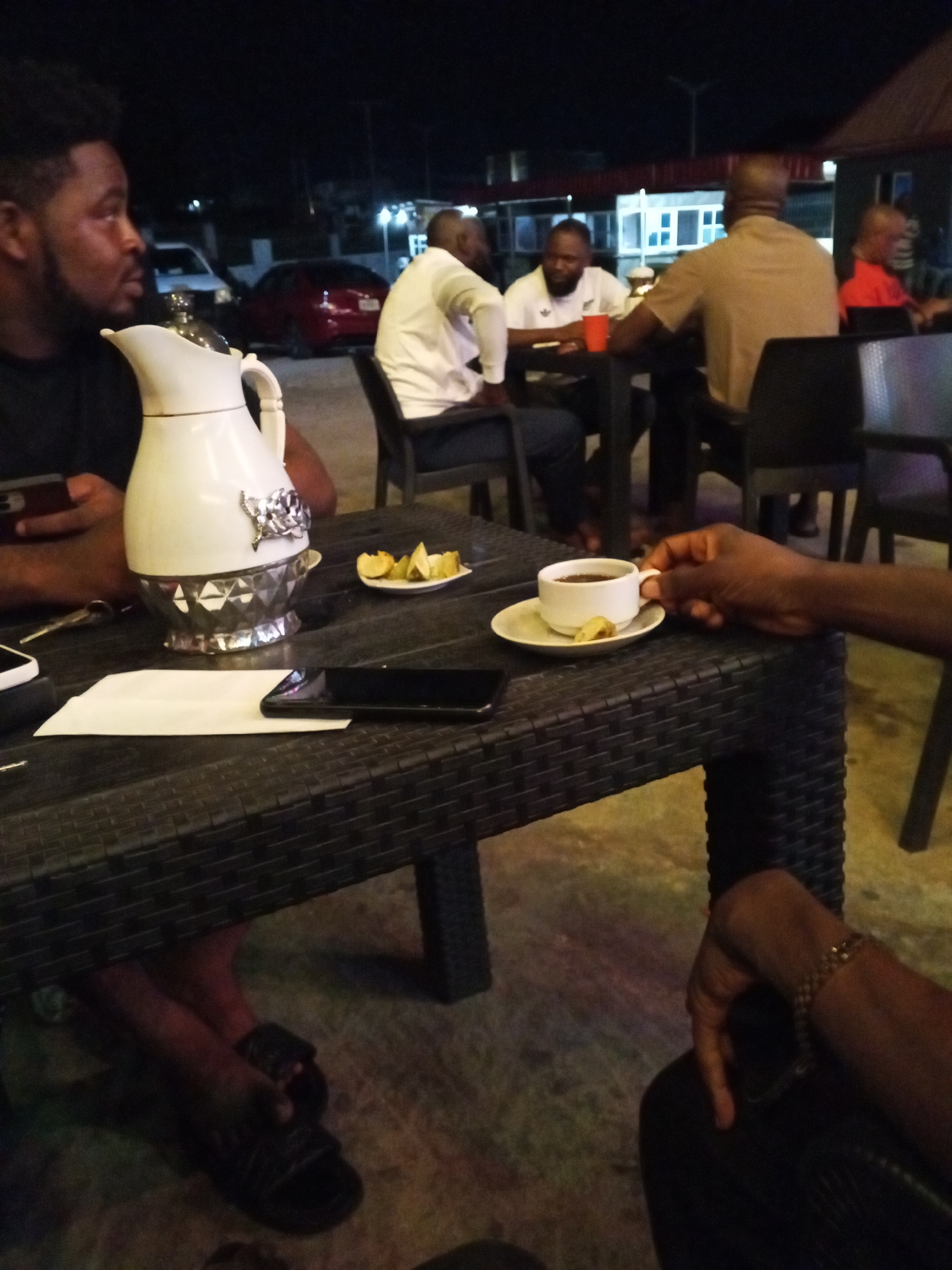 The image depicts an outdoor nighttime scene in Lokoja, Nigeria. A group of men are seated at tables, engaged in conversation and drinking beverages.
**People:**
Several men are present. One man is in the foreground, wearing a black t-shirt, and holding a smartphone. Another man is reaching for a cup. There are at least four more men at tables in the background.
**Subjects:**
The main subjects are the people, the drinks, and the objects on the tables.
**Objects:**
Objects on the table in the foreground include a white ceramic pitcher, a small plate with lime wedges, a white cup and saucer with a slice of lime, a smartphone, and a white napkin. The tables appear to be made of black wicker. There are plastic chairs.
**Actions:**
The men are drinking, talking, and looking at their phones. One is reaching for a cup.
**Interactions:**
The men are interacting with each other and with objects on the table.
**Environment:**
The scene is outdoors, likely in a restaurant or bar. The lighting is dim. In the background are a building with lights, other tables with people, and parked cars. The ground is concrete.