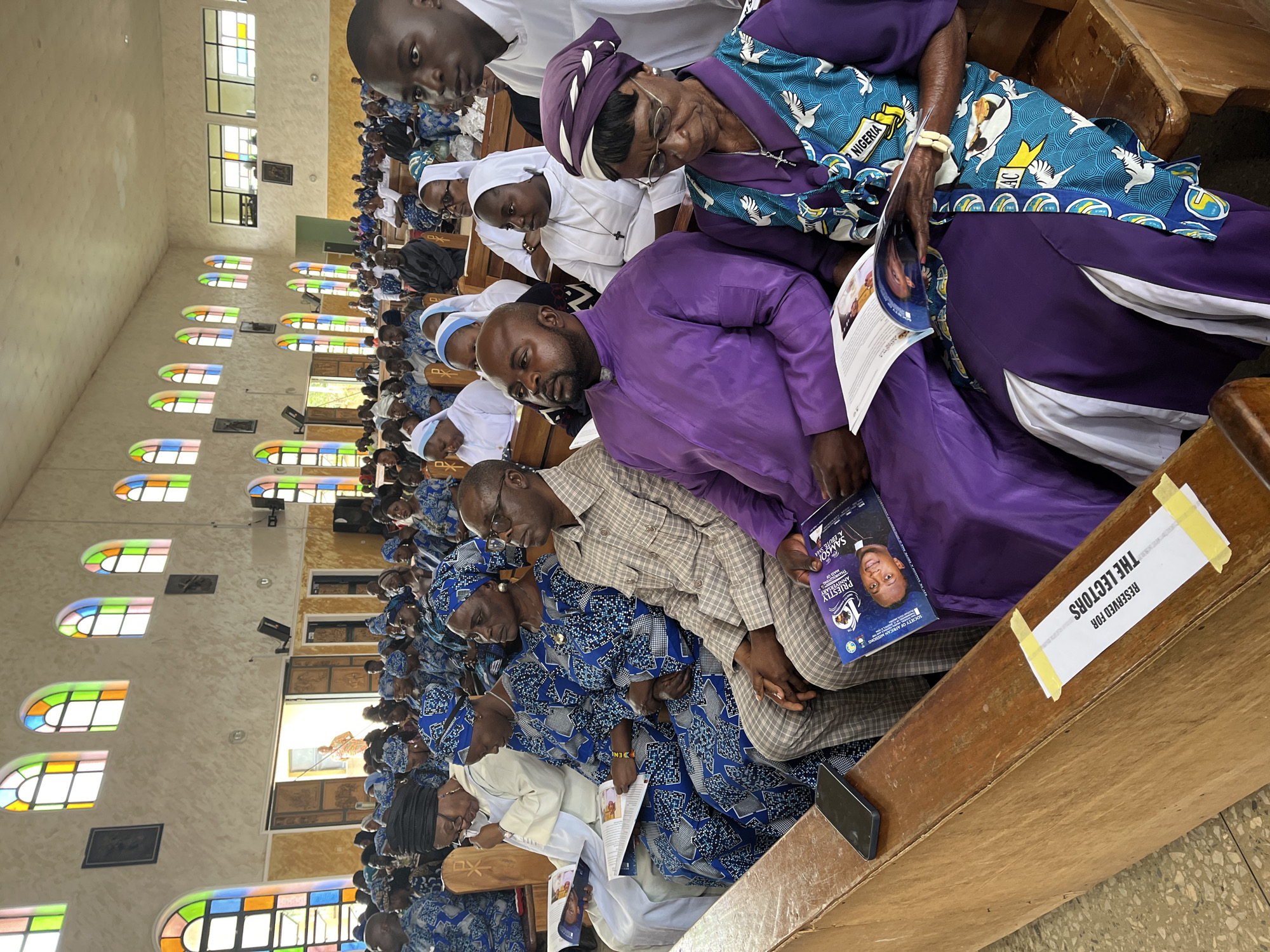 The image shows a gathering of people, primarily women, seated on wooden pews inside a church in Vom, Nigeria. Many of the women are wearing matching blue and white patterned attire, and some are wearing headscarves. Several individuals are dressed in white religious garments, suggesting a religious service or event. Some people are holding and reading booklets or programs. In the foreground, a man in a purple robe is seated, and next to him, an elderly woman in a colorful patterned outfit and headscarf is also holding a booklet. The church interior features high ceilings with arched windows filled with stained glass in vibrant colors. The walls are light-colored, and religious iconography is visible. A sign on the pew in the foreground reads "RESERVED FOR THE LECTORS." A smartphone is placed on the pew.