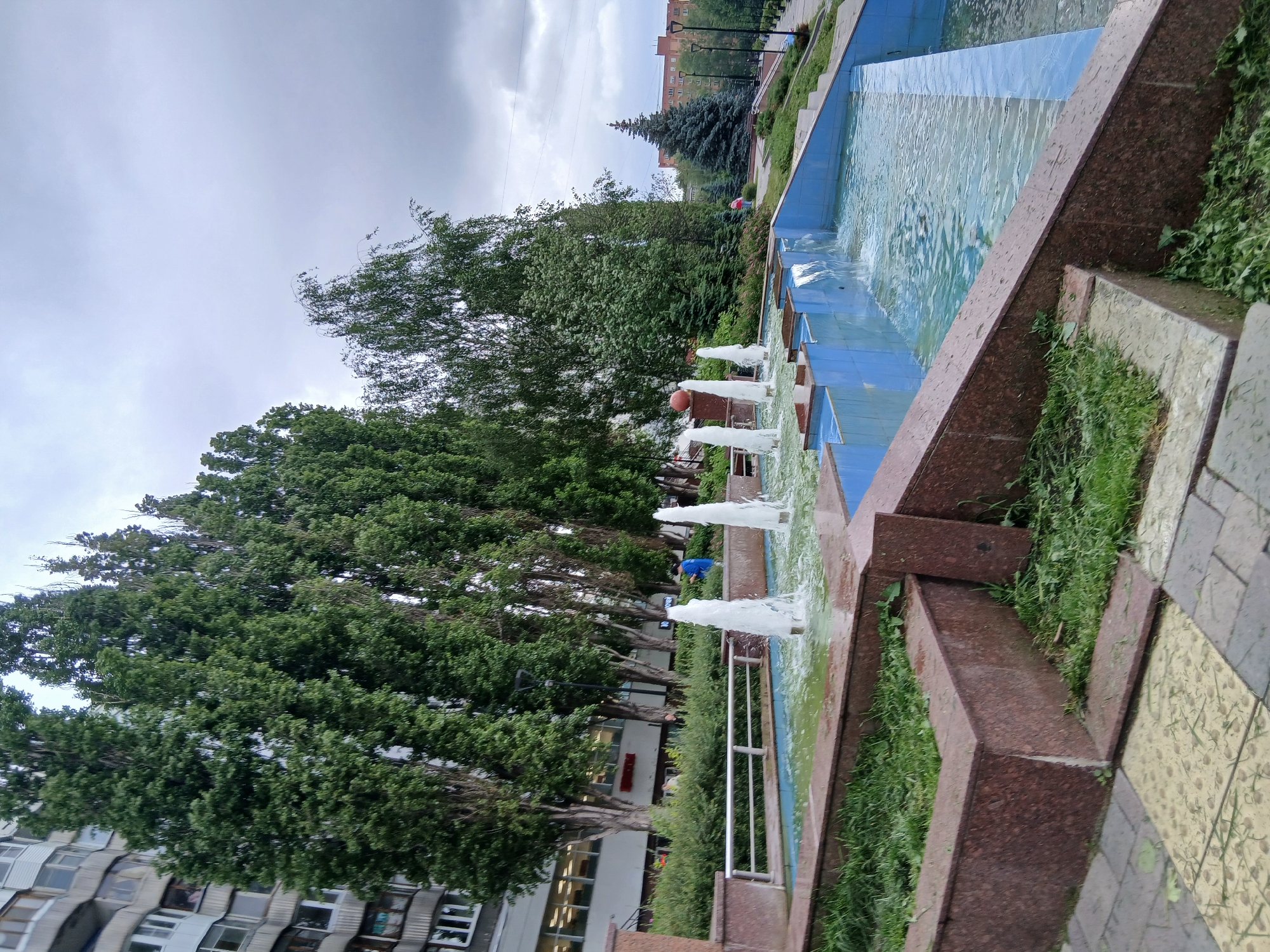 This image depicts an outdoor public space in Nizhny Novgorod, Russia, likely a park or plaza area. In the foreground, a tiered fountain with blue tiled walls and water flowing down is visible. Several jets of water are spraying upwards from the fountain's basin. The fountain is surrounded by a granite-like material, with patches of grass and debris. Behind the fountain, a row of tall, slender coniferous trees forms a dense green backdrop. To the left of the trees, a multi-story apartment building with a curved facade and numerous windows is visible, indicating an urban setting. The sky is overcast, suggesting a cloudy day. In the midground, a person in a blue shirt is standing near the fountain, and another individual in a red shirt is visible further in the background. The overall scene is one of a somewhat neglected urban public space with a fountain and surrounding greenery.