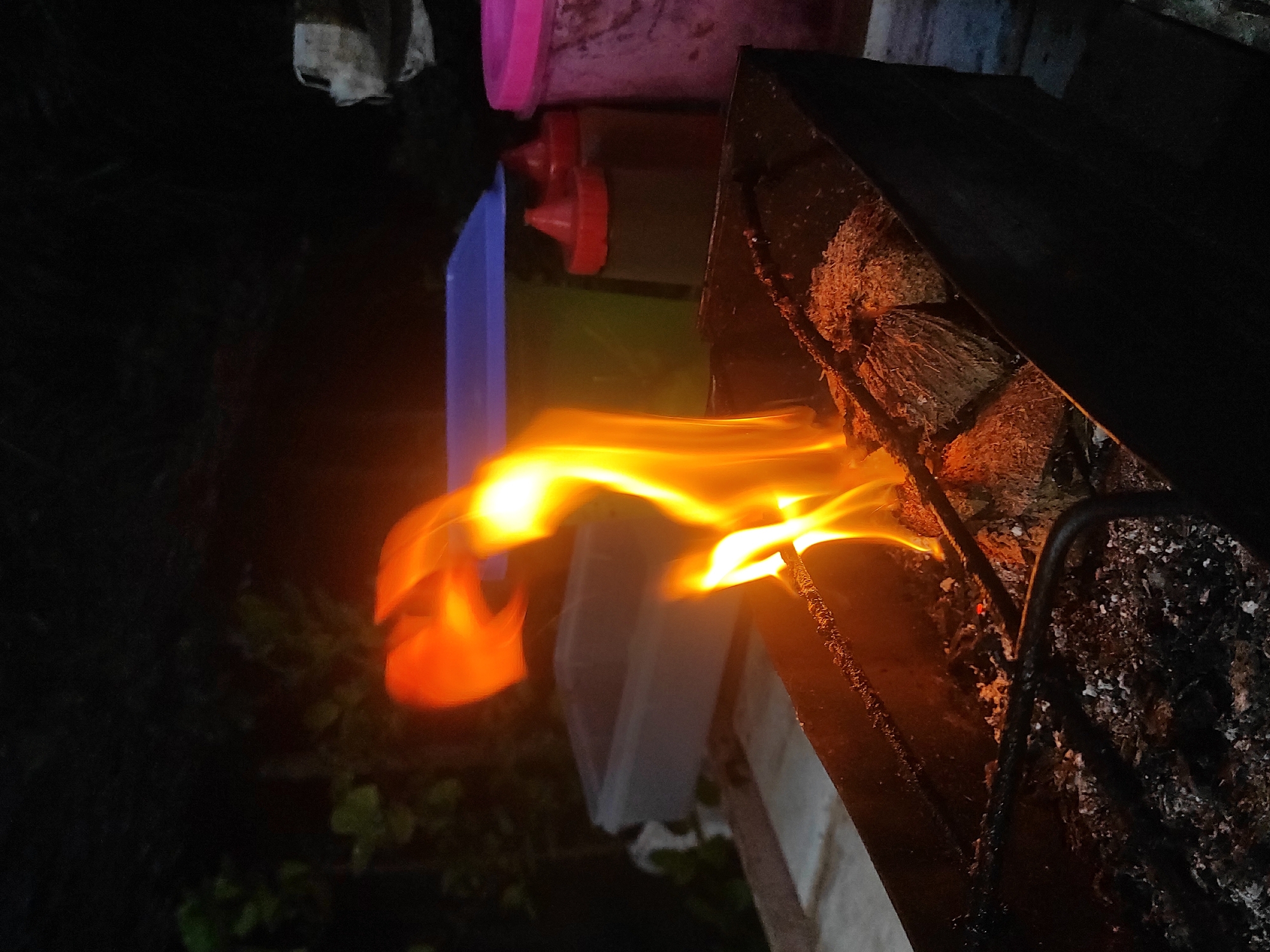 This media file captures an outdoor scene at night, focusing on a fire burning in what appears to be a barbecue grill or a similar structure. Bright, orange and yellow flames emanate from burning wood or charcoal, providing the primary light source in the dark environment.
In the foreground and center, the vibrant flames are prominent, showing dynamic movement. To the right, part of the grill structure is visible, with charcoal or burnt wood pieces and ash. Dark metal bars of the grill grate are also discernable.
Behind the fire, on a raised surface, are several objects. Two red-capped squeeze bottles, likely containing sauces or cooking oil, are clearly visible. A blue object, possibly a container or a part of the background structure, stands vertically next to them. Further back, a pink object, perhaps another container or lid, is also present. Above these items, a light-colored, crumpled cloth or plastic bag hangs in the dark background. The lower left corner shows hints of dark green foliage, suggesting natural surroundings.
The activity depicted is the burning of fuel for a fire, commonly done for grilling or warmth. The time of day is clearly night, as indicated by the intense contrast between the bright flames and the surrounding darkness. There is no visible text in the image, and no people are present. The scene suggests a preparatory phase for cooking or a casual outdoor gathering in Lhokseumawe, Indonesia, as per the provided context.