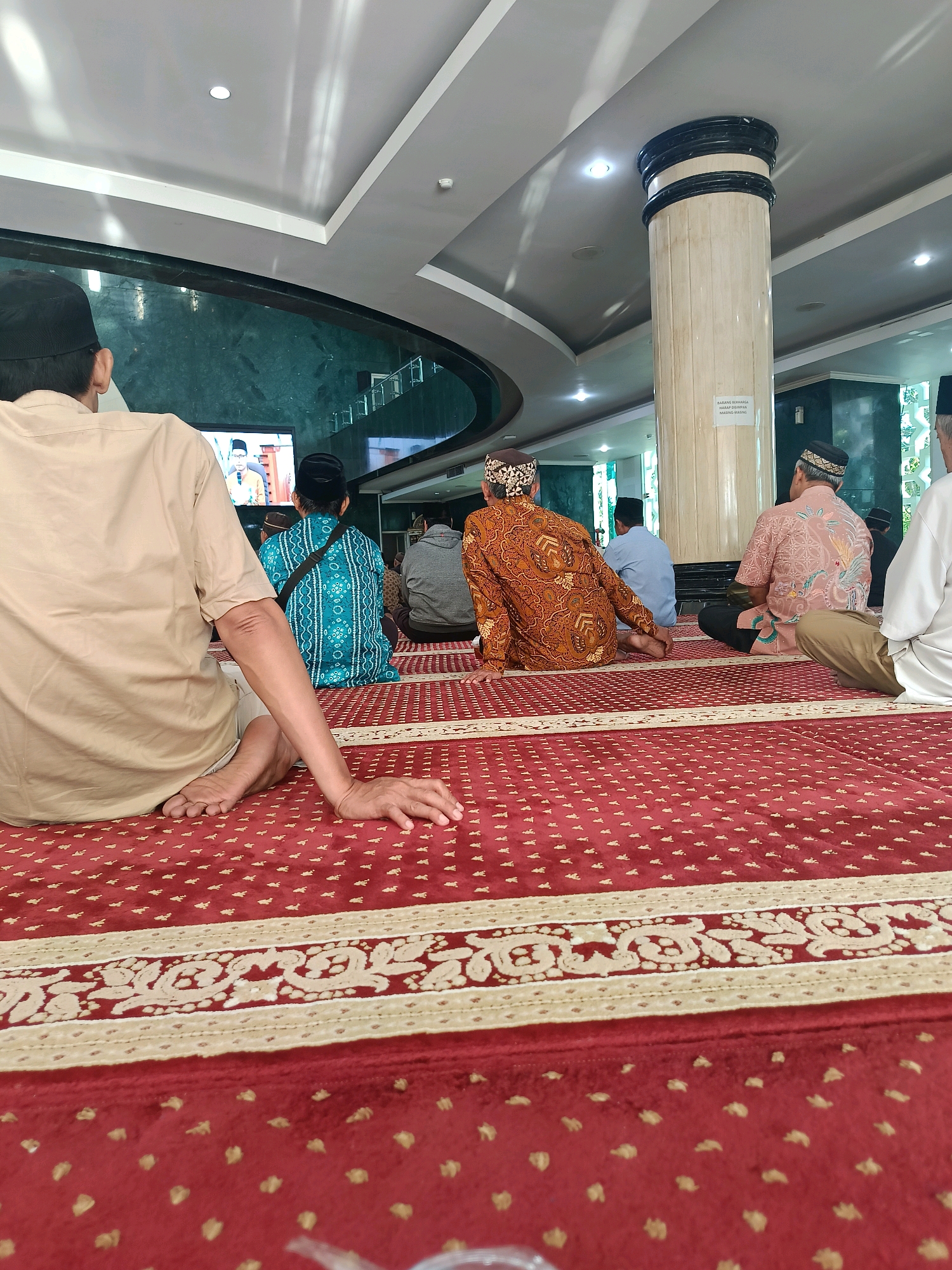 The image shows an indoor scene, likely a mosque, with several men sitting on a red carpeted floor in what appears to be a prayer or gathering. In the foreground, a man in a light brown shirt and white pants is seated with his legs crossed. His right arm rests on his leg, and his hand is relaxed. Behind him, other men are seated on the carpet, some facing a television screen visible in the background. The men are dressed in traditional attire, including peci (caps) and batik shirts. The environment includes a large pillar with text on it, a high ceiling with lighting, and green walls. The carpet has a decorative border. The image quality suggests it was taken with a mobile phone. The presence of men in prayer attire and the mosque-like setting strongly indicate this is a religious gathering. The signage on the pillar is in Indonesian, suggesting the location is in Indonesia. The specific location of Banguntapan, Indonesia, is mentioned in the prompt, which is a subdistrict in Bantul Regency, Yogyakarta Province. This context helps to interpret the cultural and religious aspects of the scene.