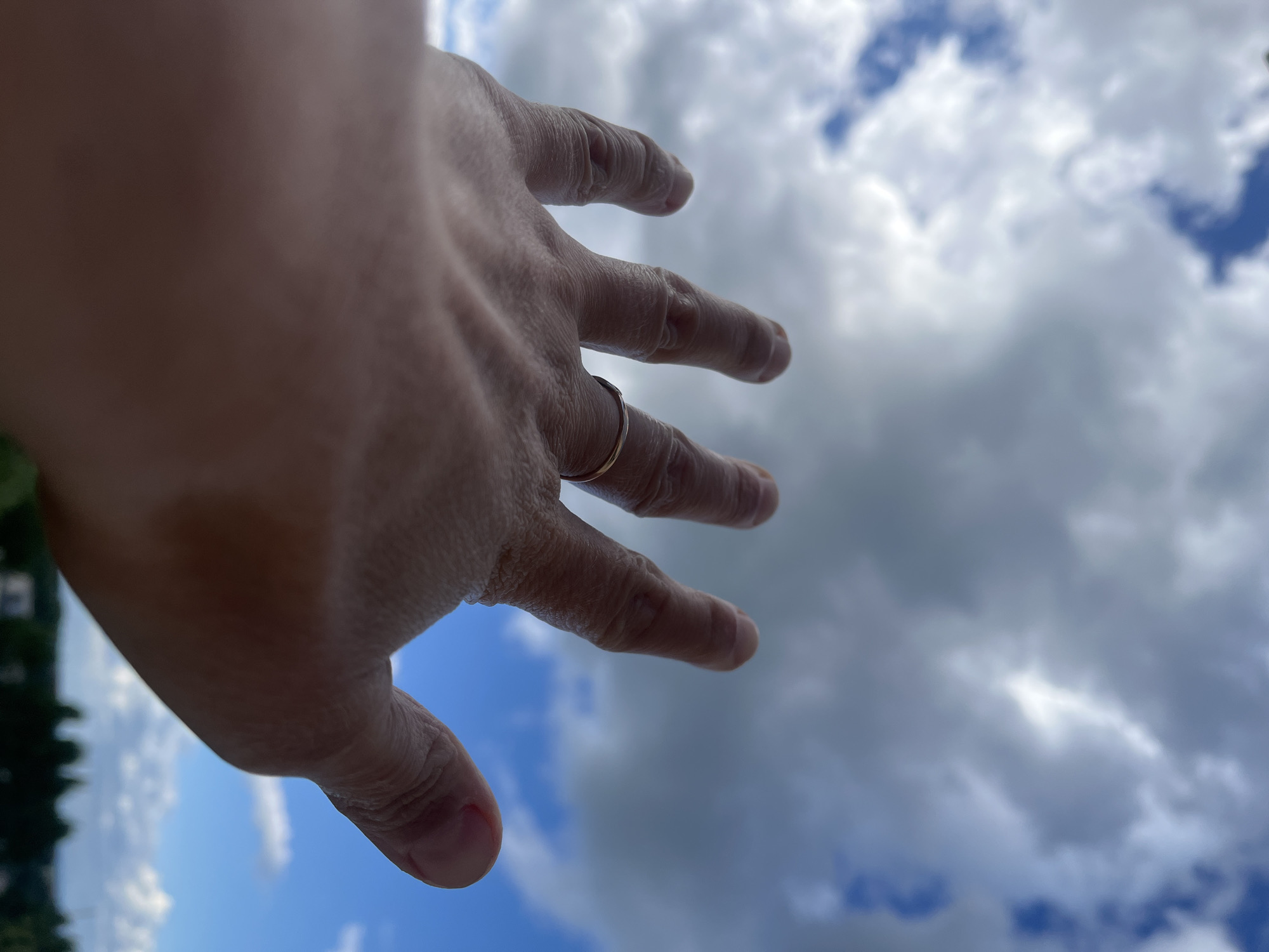 A close-up, low-angle shot focuses on a person's left hand reaching upwards towards a cloudy sky. The hand, seen from the palm side with fingers extended and slightly spread, is positioned in the lower left portion of the frame. A thin, gold-colored ring is visible on the ring finger. The sky above is filled with a mix of white and dark grey clouds, interspersed with patches of blue sky. In the far background, out of focus, a distant landscape of trees and structures can be discerned. The image appears to have been taken outdoors, under daylight conditions.