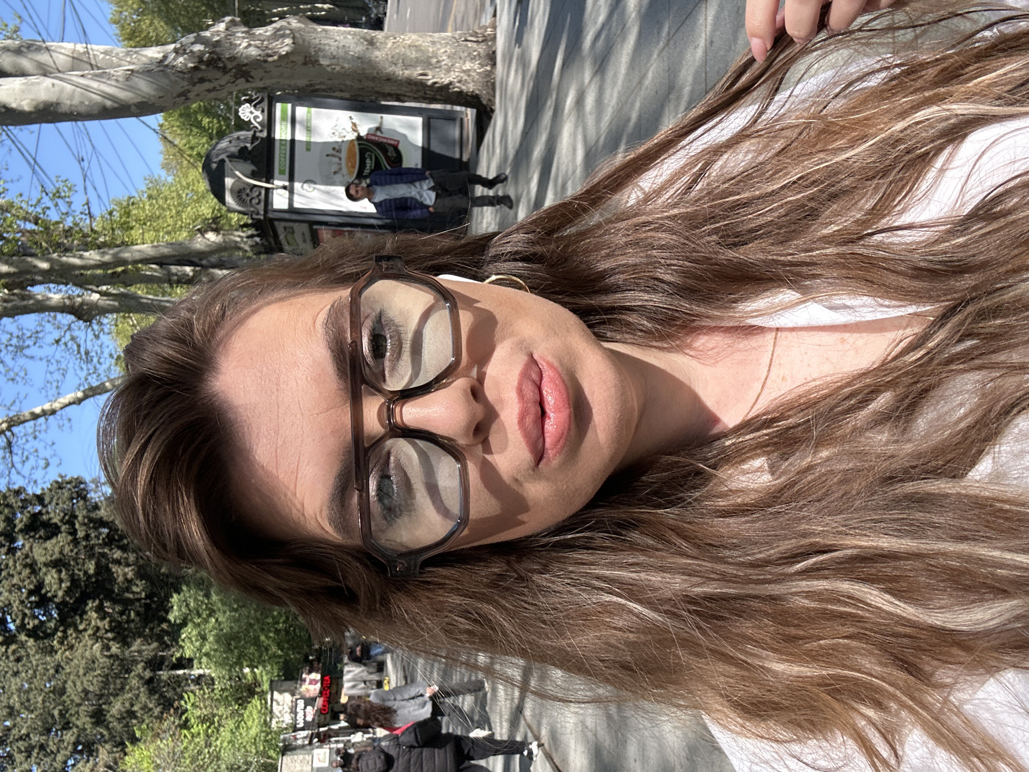 The image is a close-up, selfie-style photograph of a woman taken outdoors on a sunny day, likely in a city environment such as Tbilisi, Georgia.
The main subject is a woman, depicted from the chest up, with long, wavy brown hair featuring lighter highlights. Her hair flows over her shoulders and part of her body, which appears to be clad in a white garment. She wears large, dark-framed glasses, through which her eyes are visible. Her lips are full and have a glossy pink finish. A subtle golden chain is visible around her neck, and a white earbud is present in her right ear. Her hand is slightly visible at the top right, suggesting she is holding the camera for the selfie.
The background reveals an urban setting under a clear blue sky. Large trees with light-colored, possibly sycamore, bark and green foliage are prominent. In the mid-ground, there is a dark kiosk or street vendor stand. Visible text on the kiosk includes "COFFEE & MORE", "AMERICANO", and "TEMPO". Several people are seen in the background, walking along a sidewalk or street, contributing to a lively city atmosphere. One man in a blue jacket is standing near the kiosk.
The photograph captures a casual moment, with the woman taking a picture of herself against the backdrop of a city street. The strong sunlight and clear blue sky indicate it is daytime. The overall scene suggests a pleasant day for an outdoor stroll or activity in an urban setting.